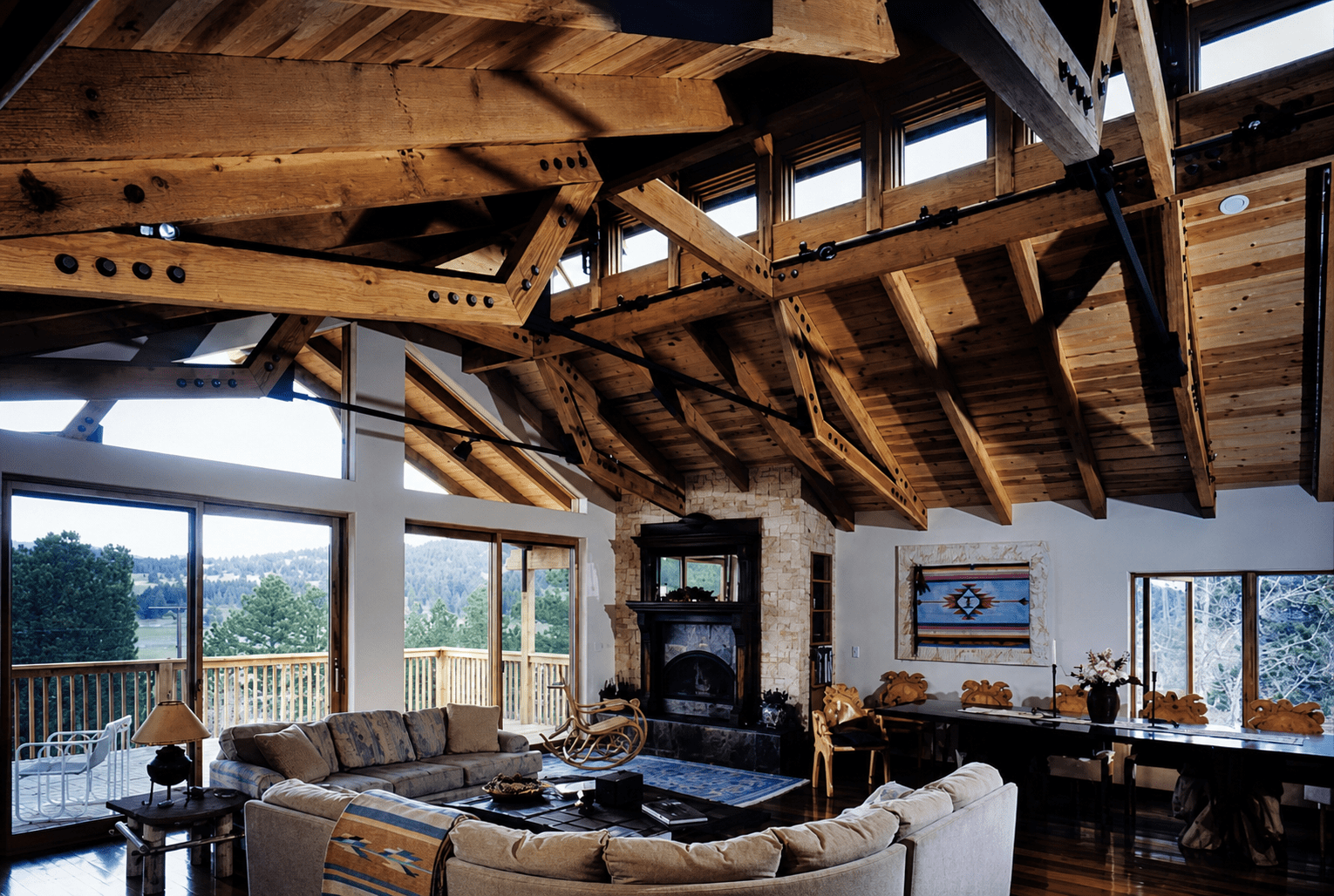 Living room with a vaulted wooden ceiling, large windows, a stone fireplace, and a mix of traditional and rustic furniture including sofas and a rocking chair, overlooking a wooded landscape.