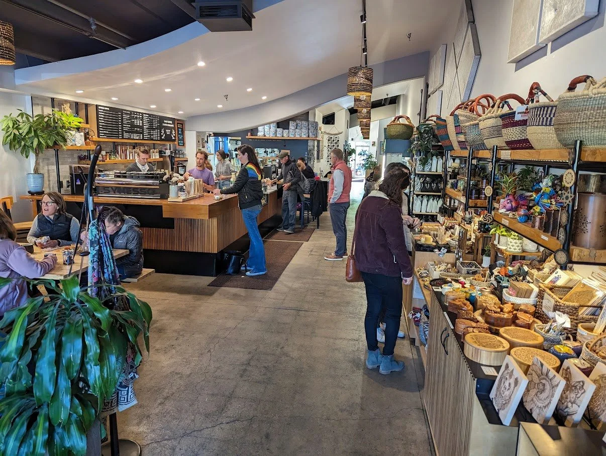 Interior of a cozy coffee shop with a wooden counter, baristas preparing drinks, customers ordering and sitting, and a display of handmade crafts and baskets on shelves along the wall.