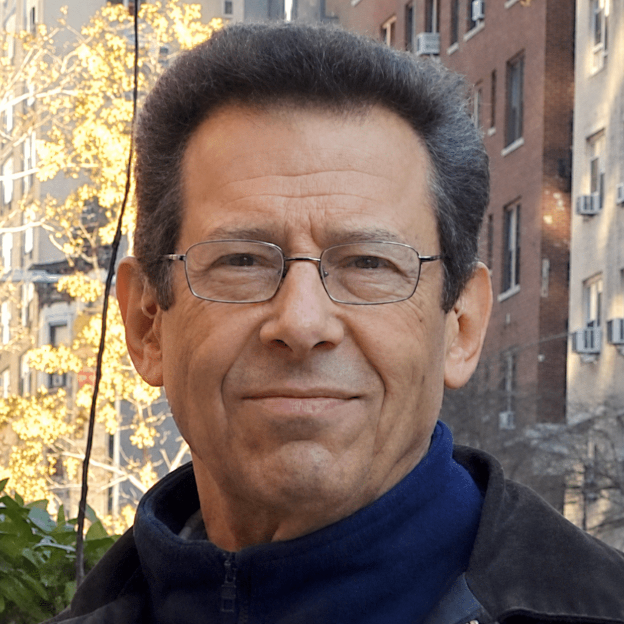 A middle-aged man with glasses smiling outdoors in a city setting with apartment buildings and yellow-leaved trees in the background.