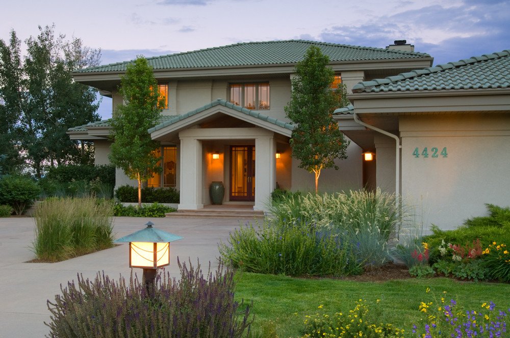 A two-story house with a green tiled roof, beige walls, and a front porch. There are two small trees and landscaped bushes in the front yard, with a garden bed full of colorful flowers. The house number 4424 is visible on the side of the house. A pathway leads to the front door, which is illuminated by wall-mounted lights.