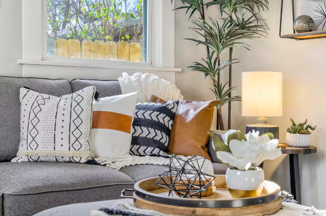 Living room with gray sofa, decorative pillows, a wooden tray with a geometric sculpture and a white flower in a white and gold vase, a side table with a lamp, a potted plant, and a window with greenery outside.