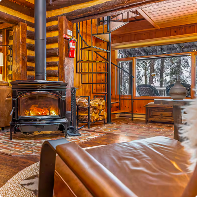 Cozy log cabin interior with a wood stove, stacked firewood, spiral staircase, and large window with outdoor trees.