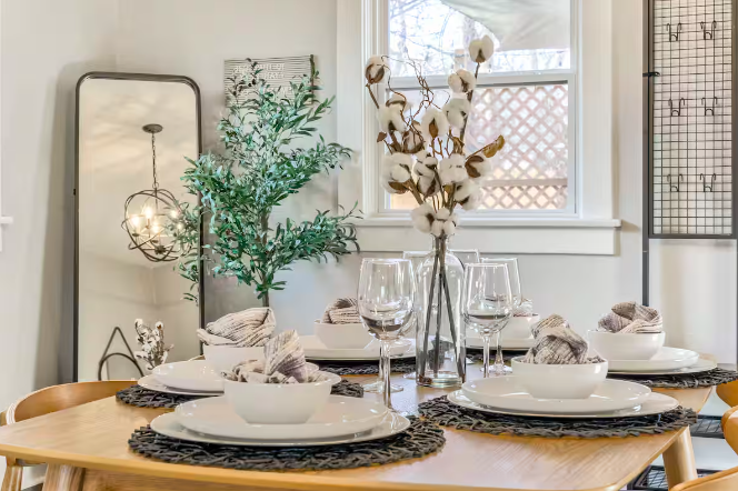 A dining table set with white plates, glassware, and napkins, decorated with a tall vase of cotton branches, in a bright room with a window, a mirror, and indoor plants.