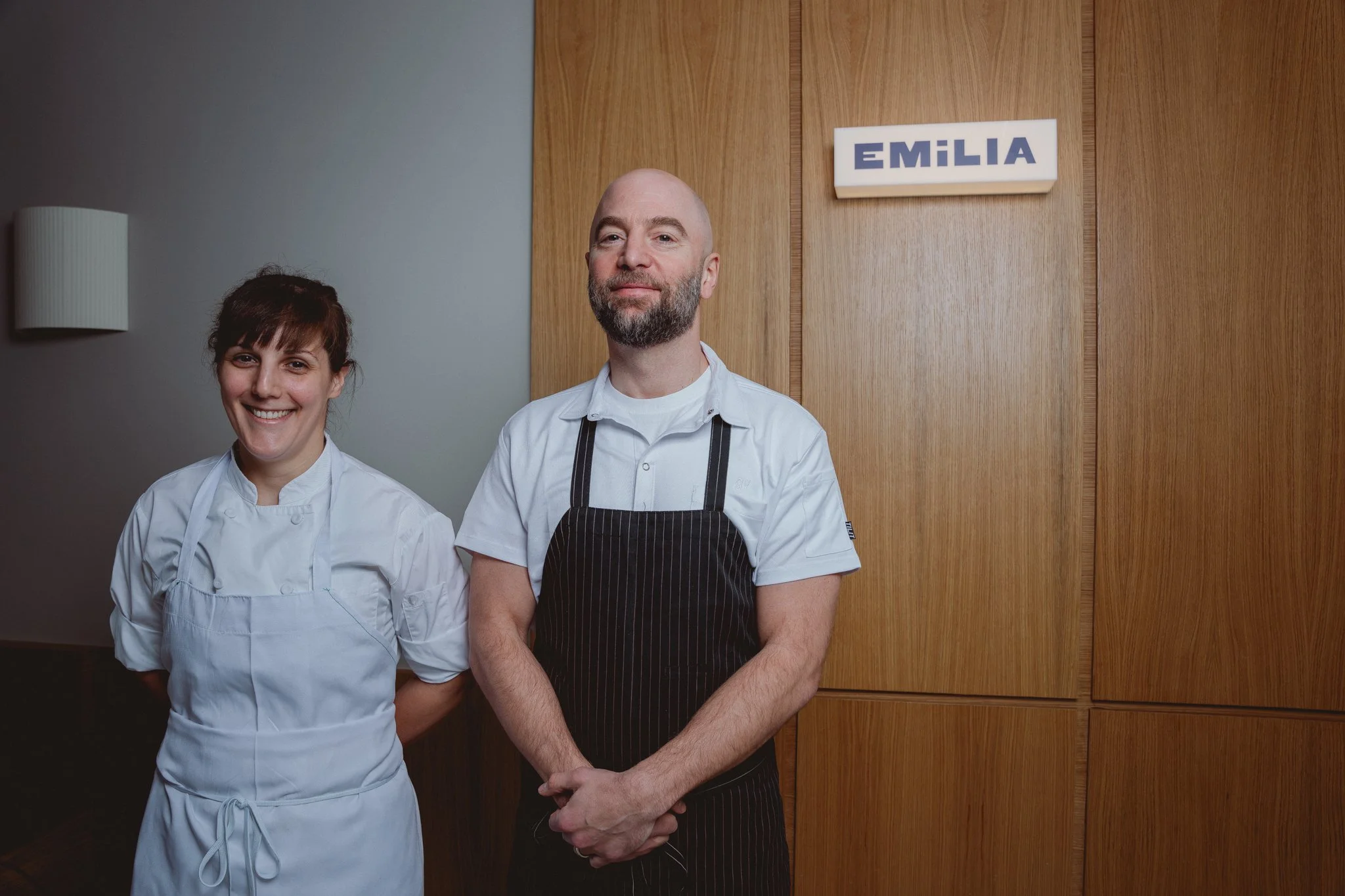 Two chefs, a woman with dark hair and a man with a beard, standing side by side in a kitchen. The woman is smiling, wearing a white chef's coat, and the man is looking at the camera, wearing a white shirt with a black striped apron. Behind them, ther