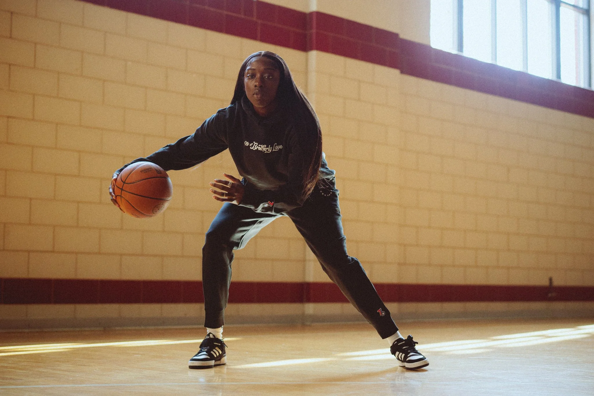 A young woman playing basketball in an indoor gymnasium, dribbling the ball with her right hand, wearing a black hoodie, black pants, and athletic shoes.