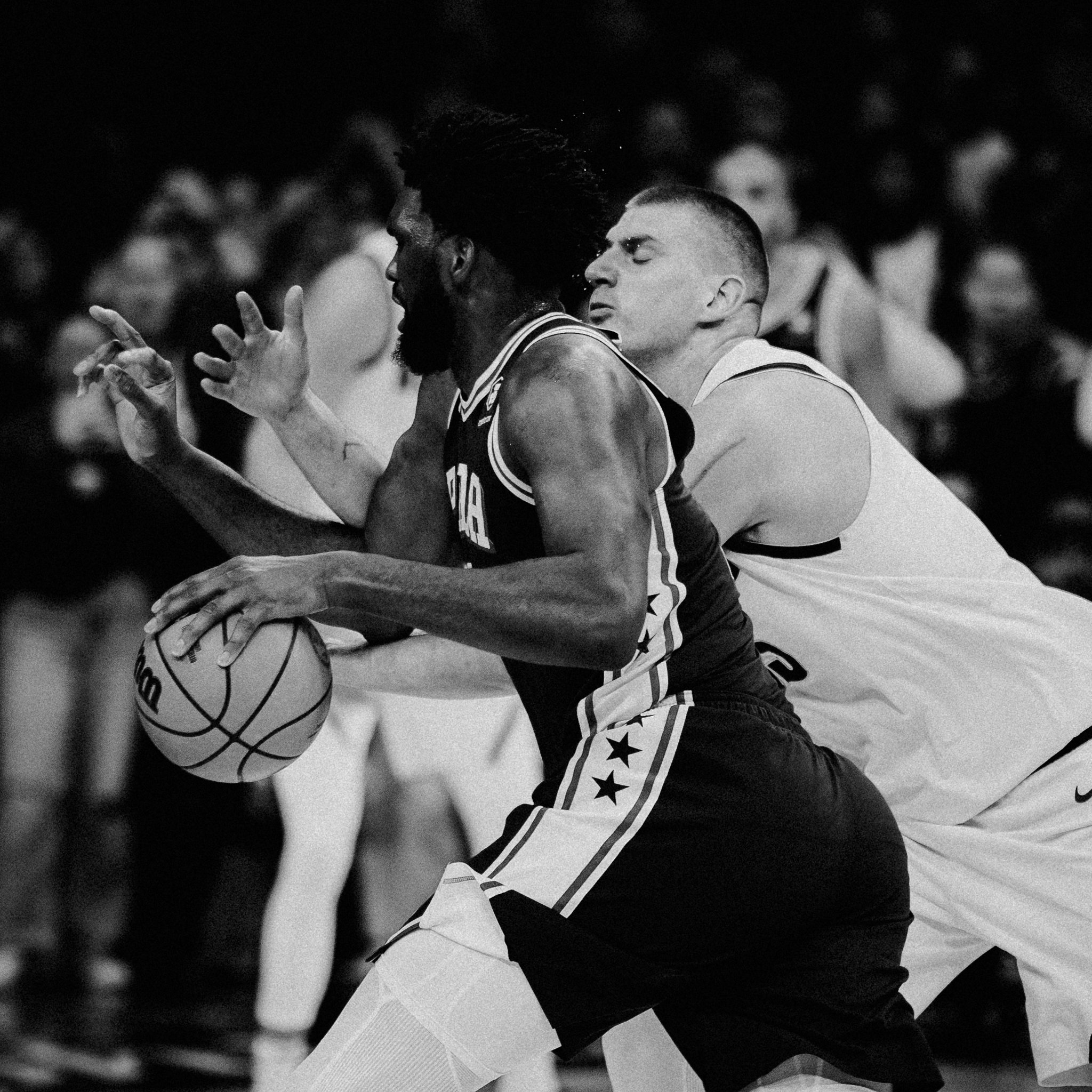 Two basketball players engaged in a game, one with dark skin and dreadlocks, the other with light skin and a short haircut, fighting for the ball amidst a crowd.
