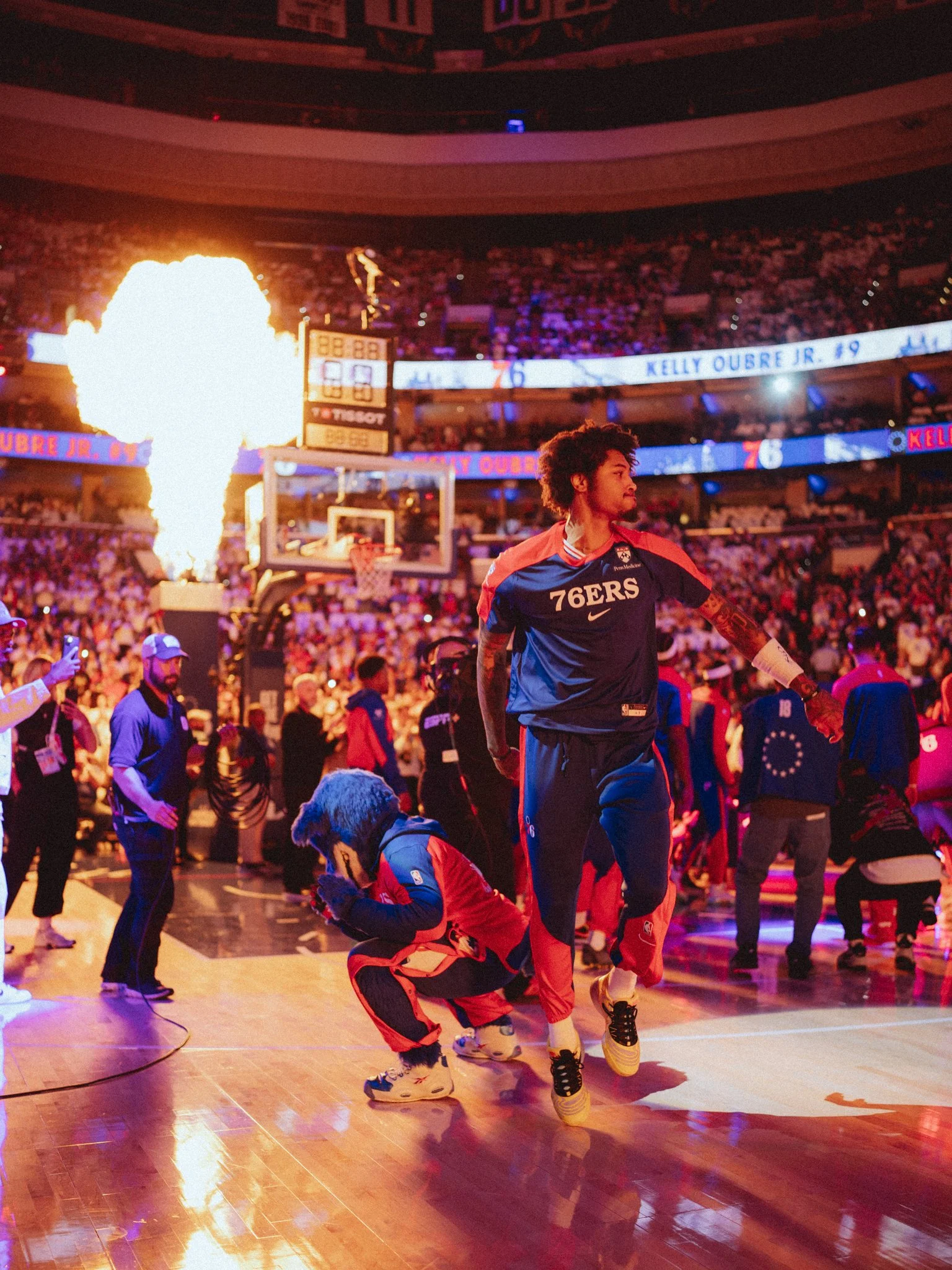 A basketball player in a Philadelphia 76ers jersey stands on the court with a fiery explosion in the background, while the audience watches from the stands. The player is looking to the side as others and security personnel are nearby.