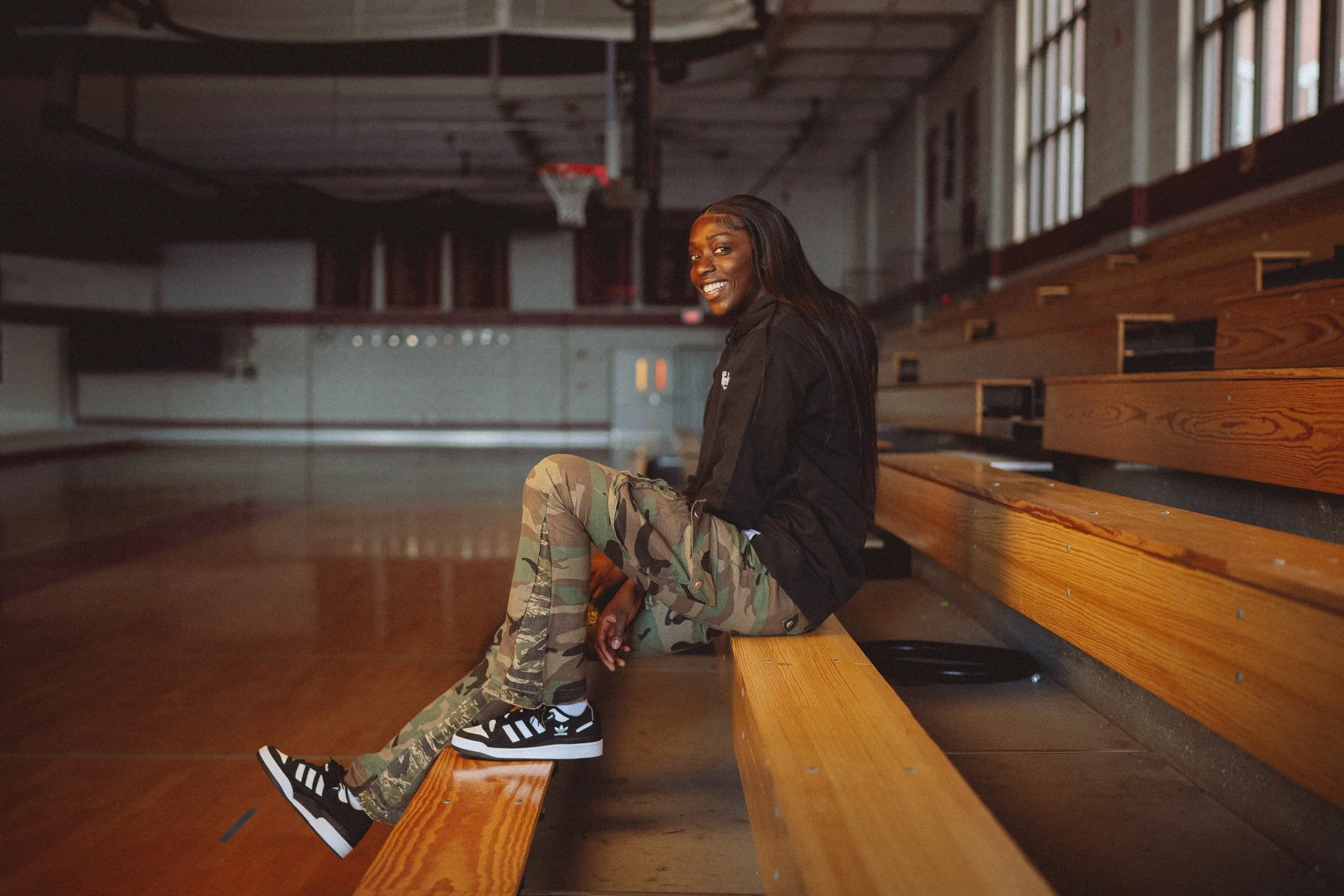 A young woman with dark hair smiling, sitting on a wooden gym bench inside a gymnasium with a basketball hoop in the background, wearing a black jacket, camouflage pants, and black and white sneakers.