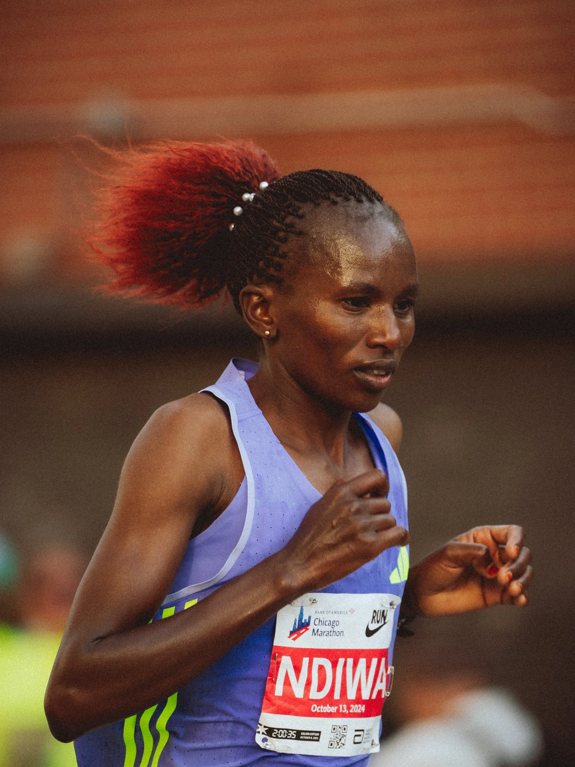 Close-up of a woman running in a marathon, wearing a purple athletic tank top, with a race bib that reads 'INDIANA' and 'October 13, 2024'. She has dark braided hair with a red extension, decorated with white beads, and appears focused while running.