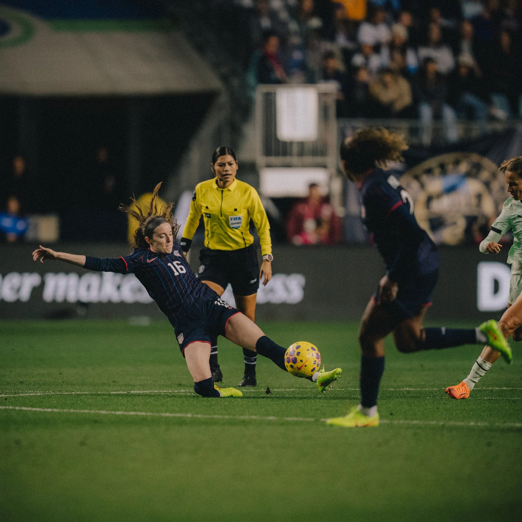 Female soccer players competing for the ball during a match with a female referee observing, on a green field with a crowd of spectators in the background.