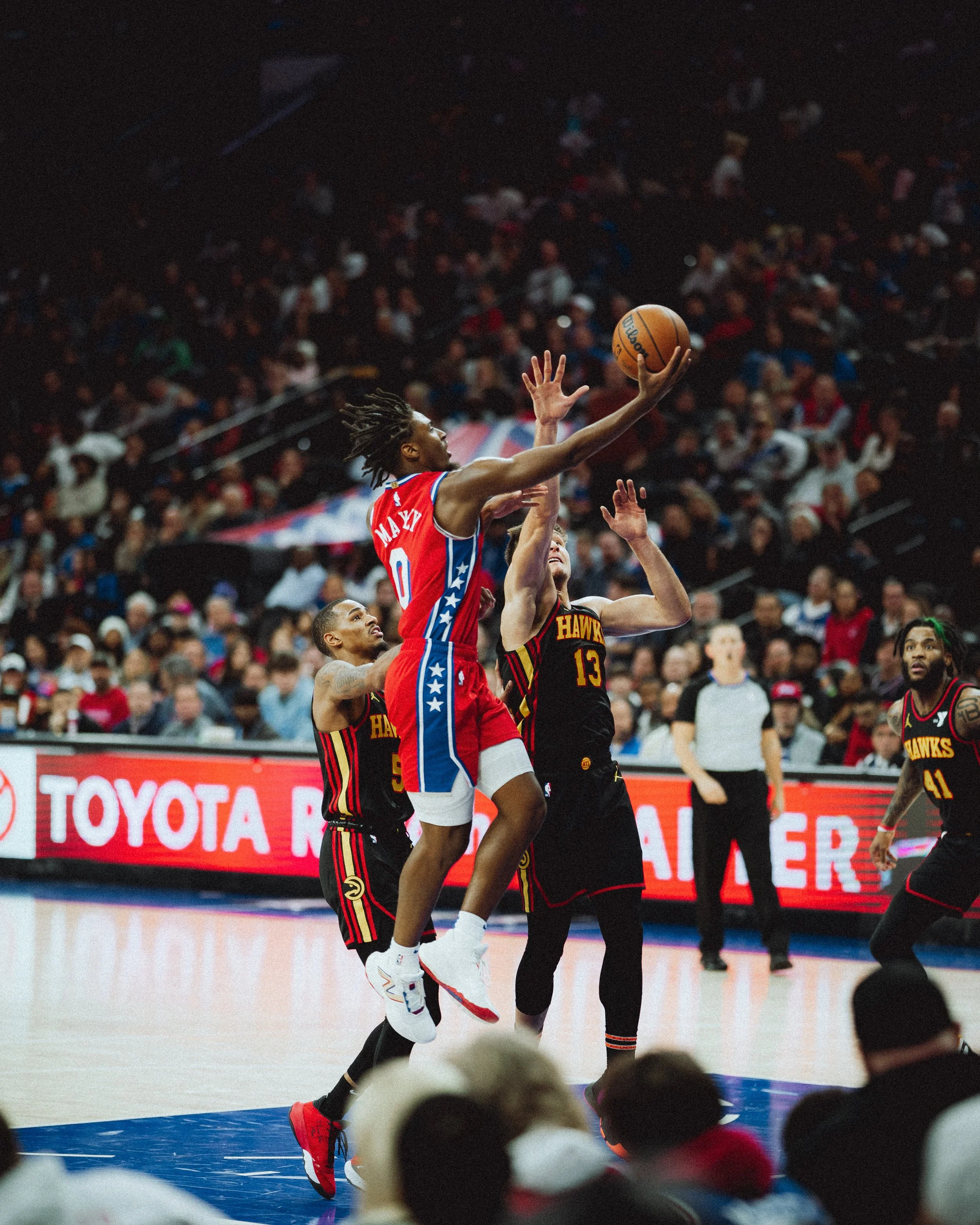A basketball player in a red and blue jersey with the number 0 jumps to shoot the ball while another defender in a black and red jersey with the number 13 attempts to block the shot during a game in an arena filled with spectators.