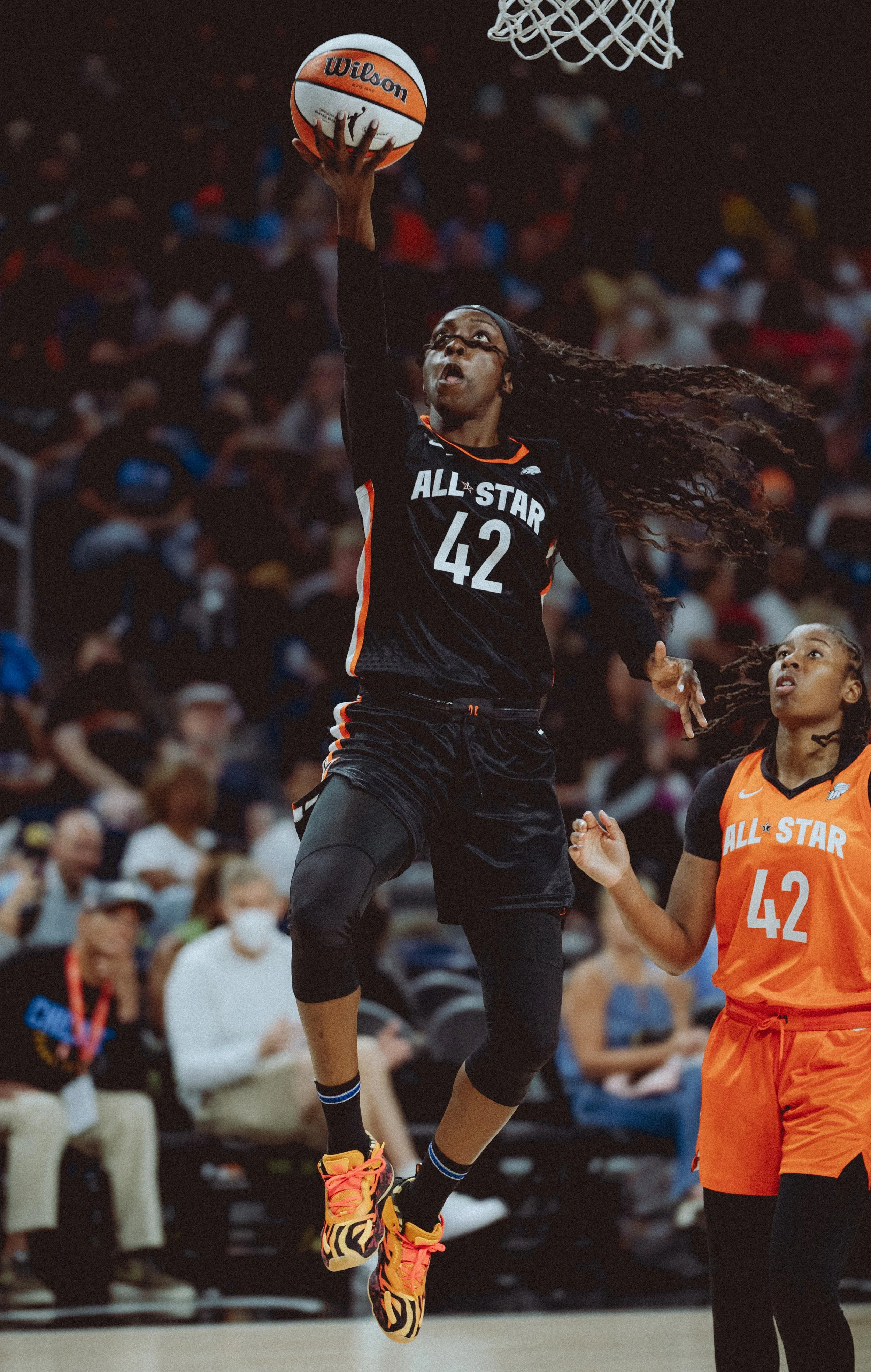 A woman in black sports attire with number 42 and glasses is jumping to make a shot during a basketball game. She is holding a basketball labeled Wilson, with her hair flowing as she leaps near the basket. Another player in an orange uniform with the