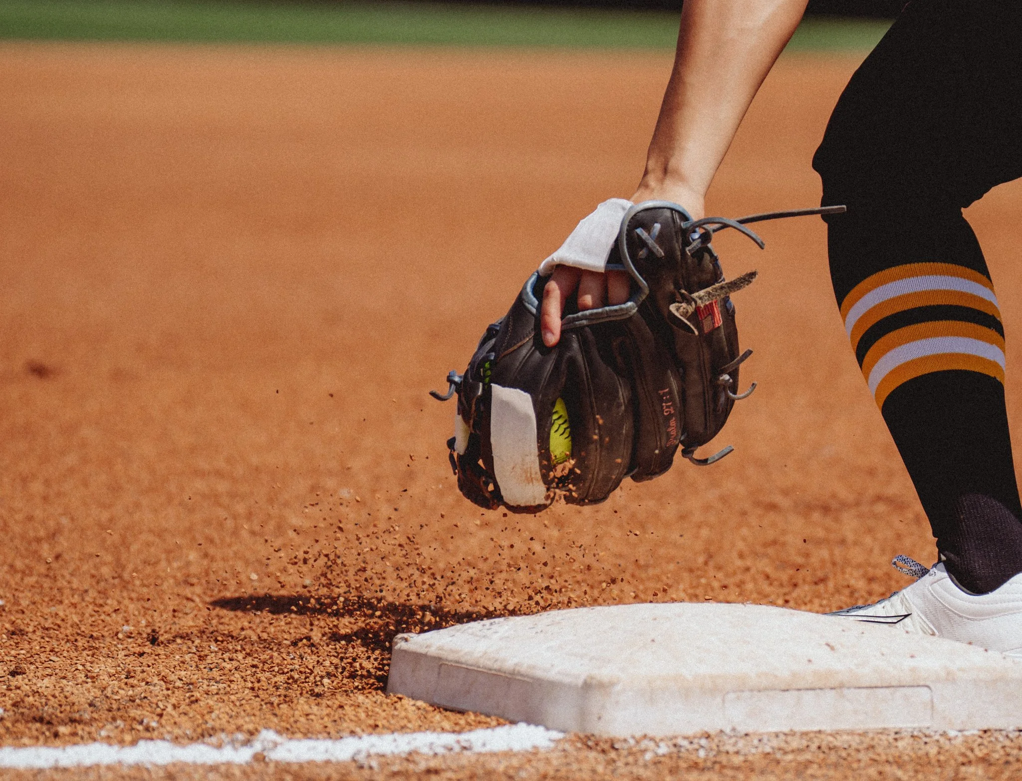 Close-up of a baseball pitcher’s foot on the rubber, with a baseball glove held in the hand, on a dirt mound next to a white line on a baseball field.