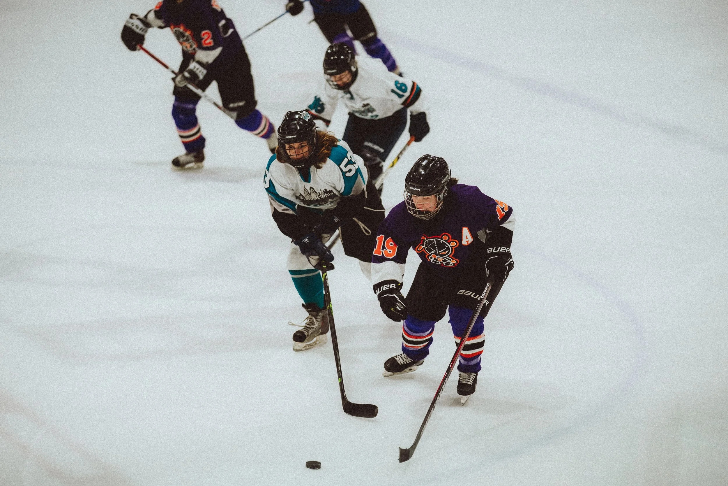 Four young girls playing ice hockey on an ice rink, wearing helmets and hockey gear, skating and competing for the puck.
