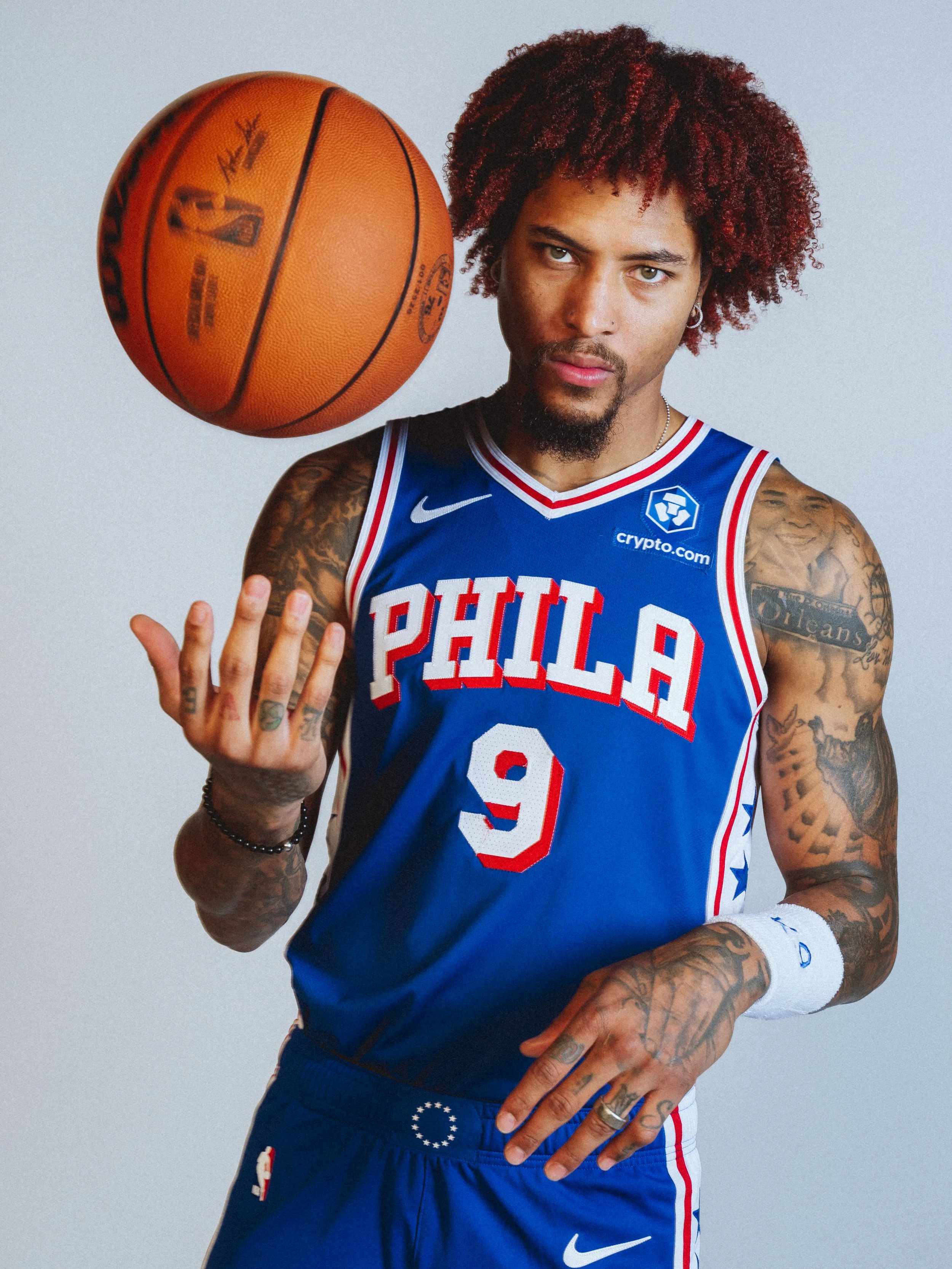 NBA Player,  Kelly Oubre with tattoos on his arms, wearing a blue Philadelphia 76ers jersey, spins a basketball on his finger, with a neutral gray background.