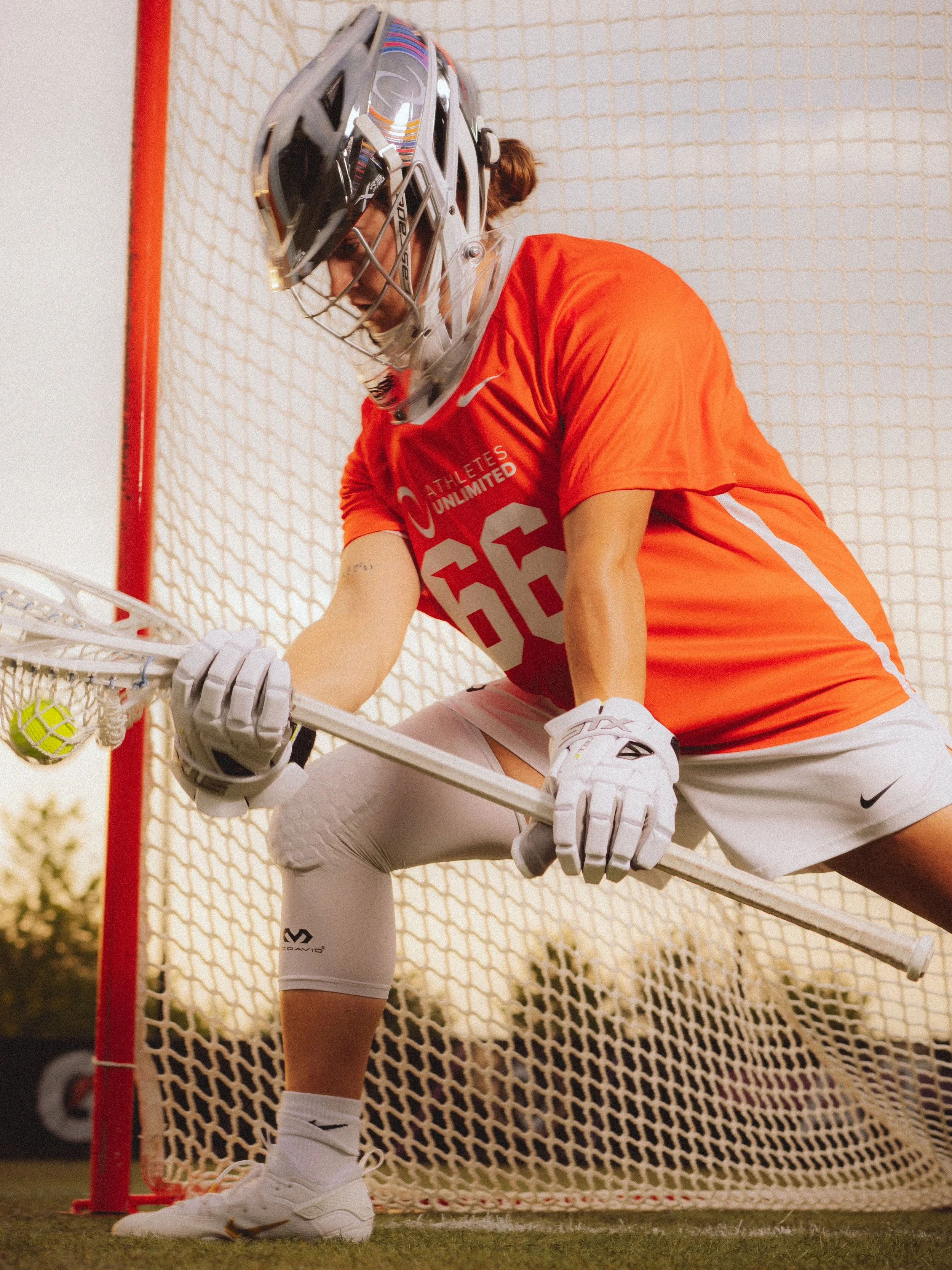 A female field hockey player wearing a red jersey and white shorts, preparing to defend near the goal, with a goal net behind her and a field hockey ball on the ground.