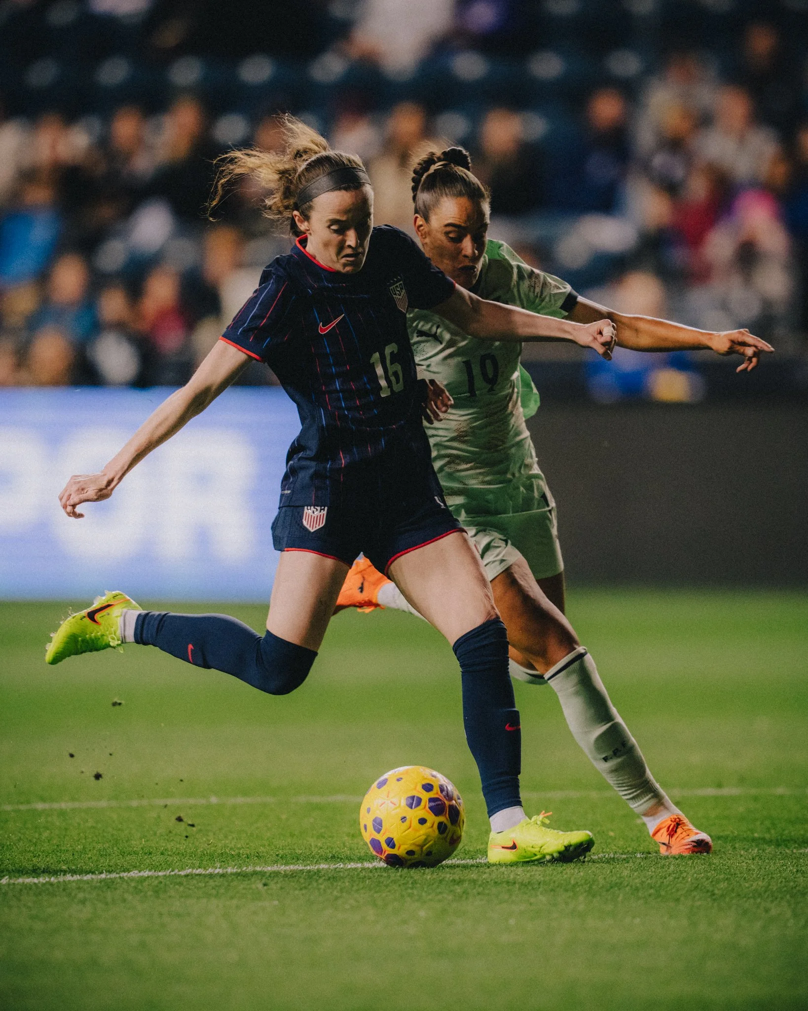 Two female soccer players compete for the ball on the field during a game. One player from the U.S. women's team, wearing a navy jersey with red and white accents, number 16, is focused on the ball. The other player, wearing a green uniform with the 