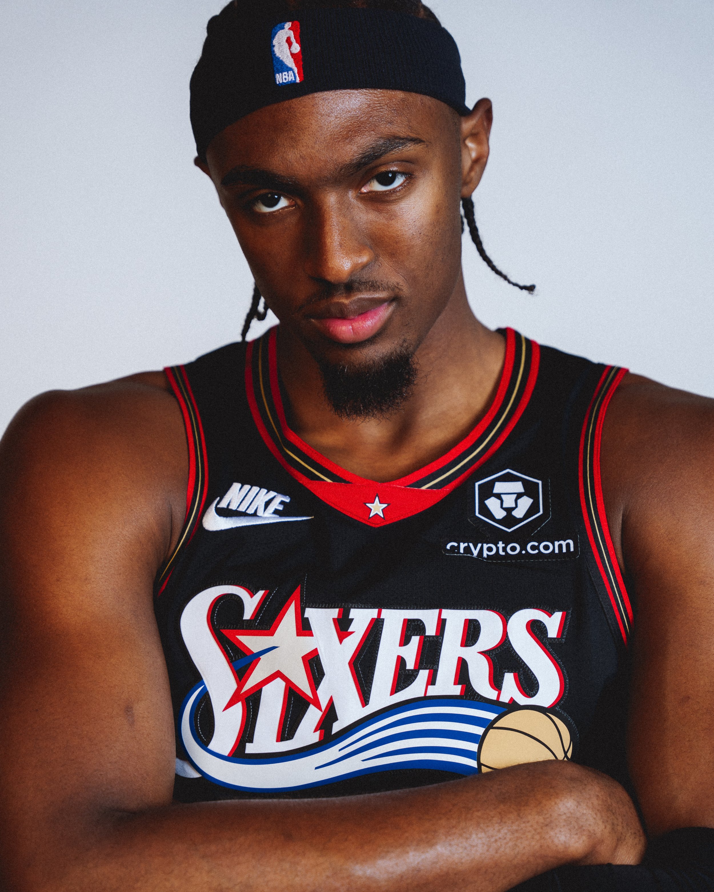 NBA Player, Tyrese Maxey wearing a black basketball jersey and a black NBA headband, with a tattoo on his right arm, against a plain background.