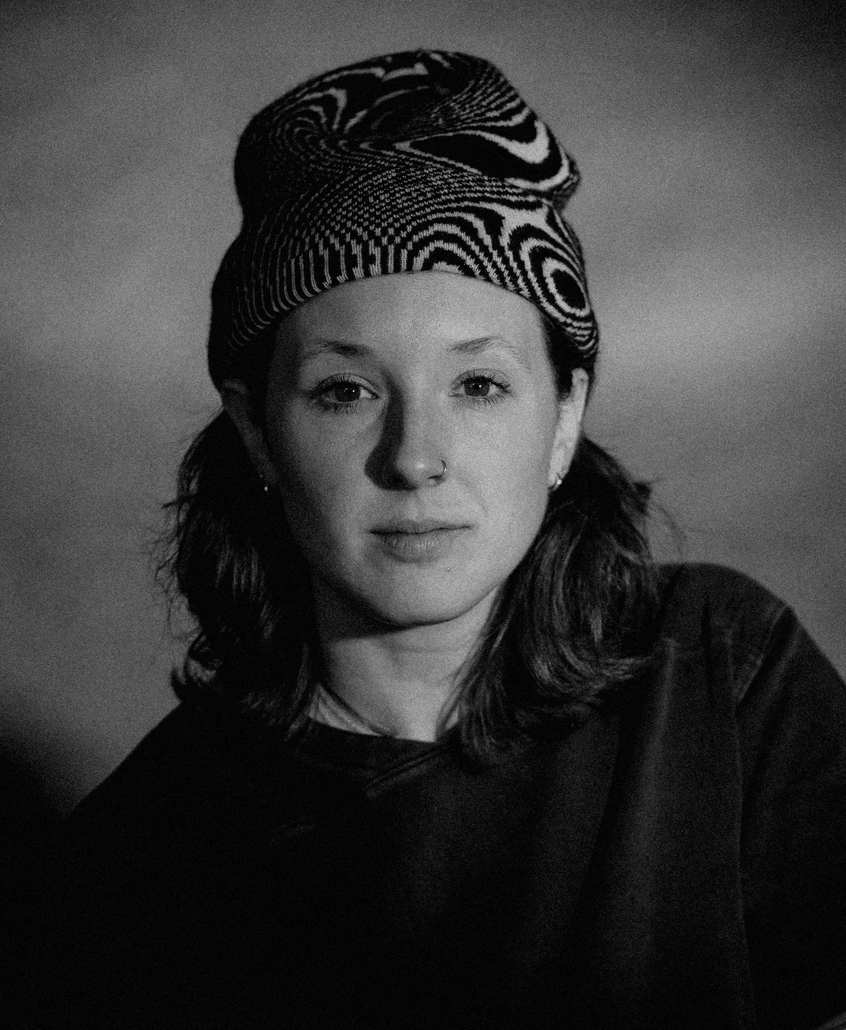 Black and white portrait of a woman with a patterned headscarf covering her hair, with a nose ring, looking directly at the camera.