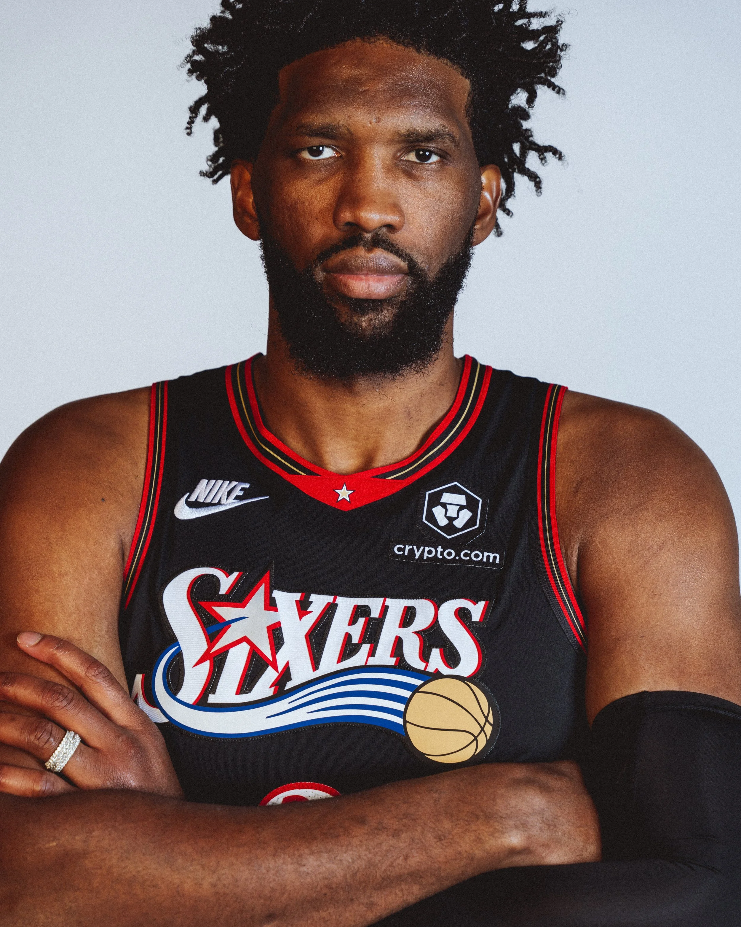 NBA Player, Joel Embiid stands with arms crossed, wearing a black jersey with red and white accents, featuring the Philadelphia 76ers logo and sponsor logos.