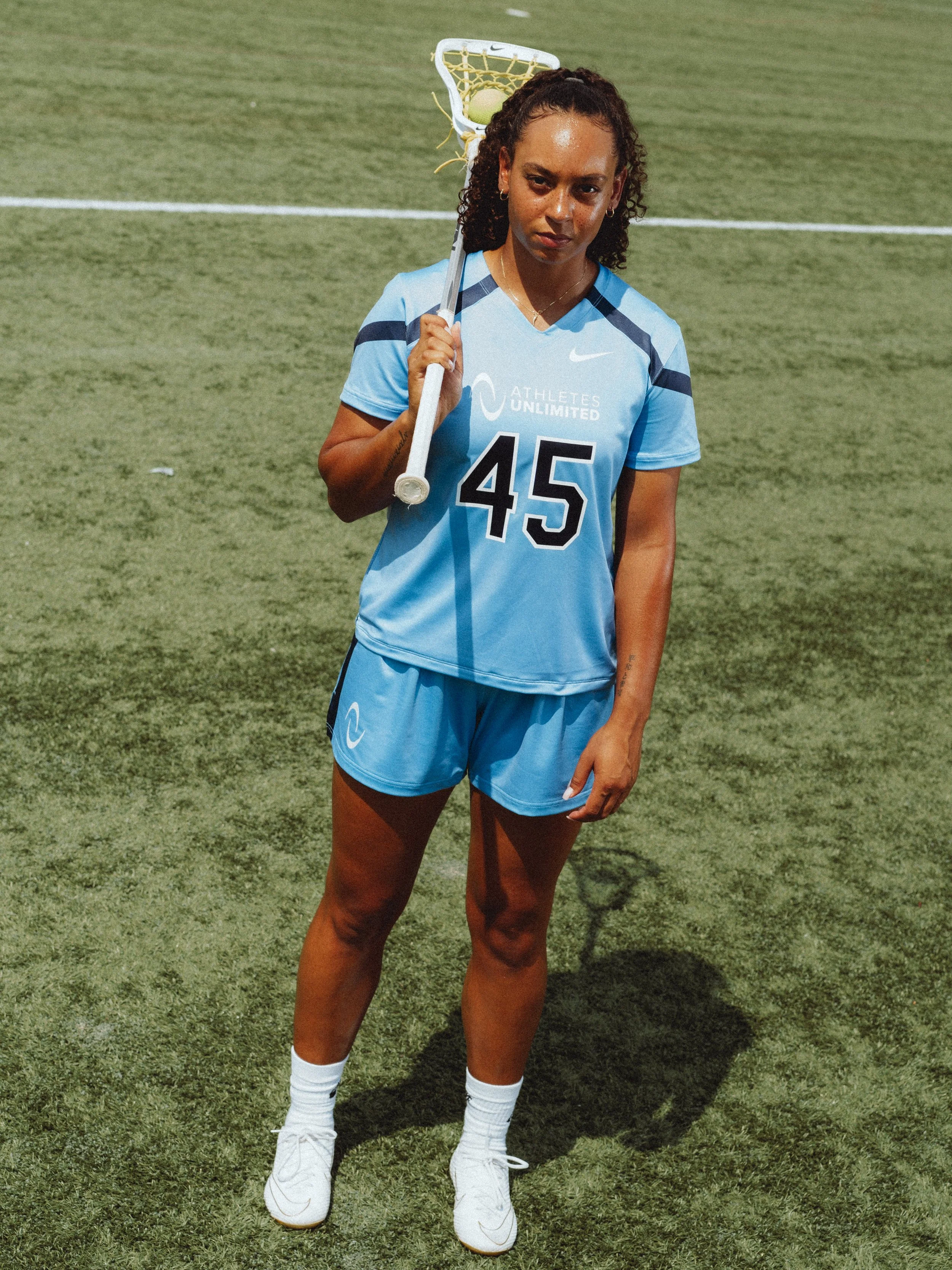 A female lacrosse player in a light blue uniform holding a lacrosse stick with a ball in the net, standing on a grassy field.
