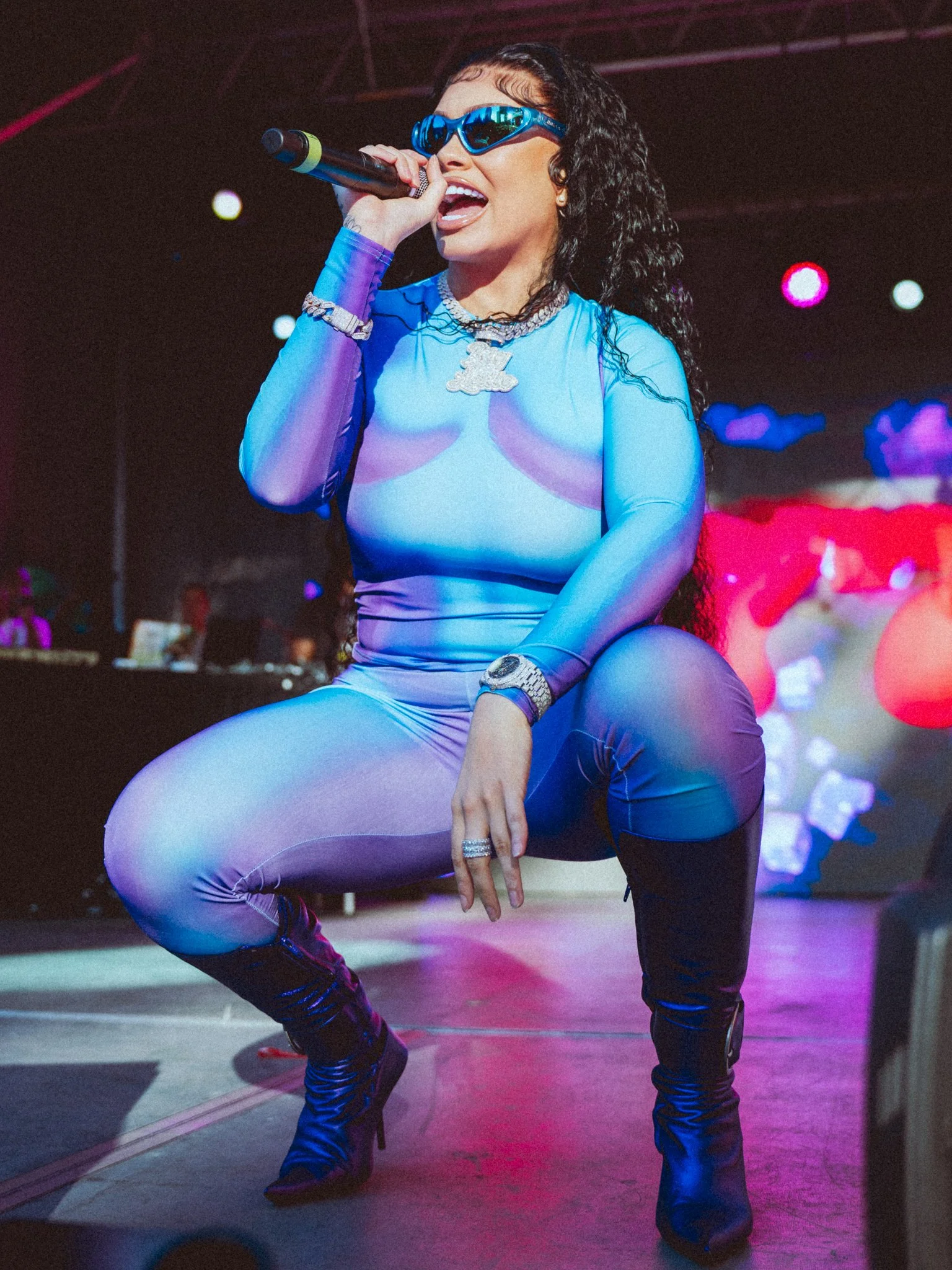 A performer singing on stage while wearing blue sunglasses, a shiny matching blue outfit, and black thigh-high boots with high heels, holding a microphone, with colorful stage lights in the background.