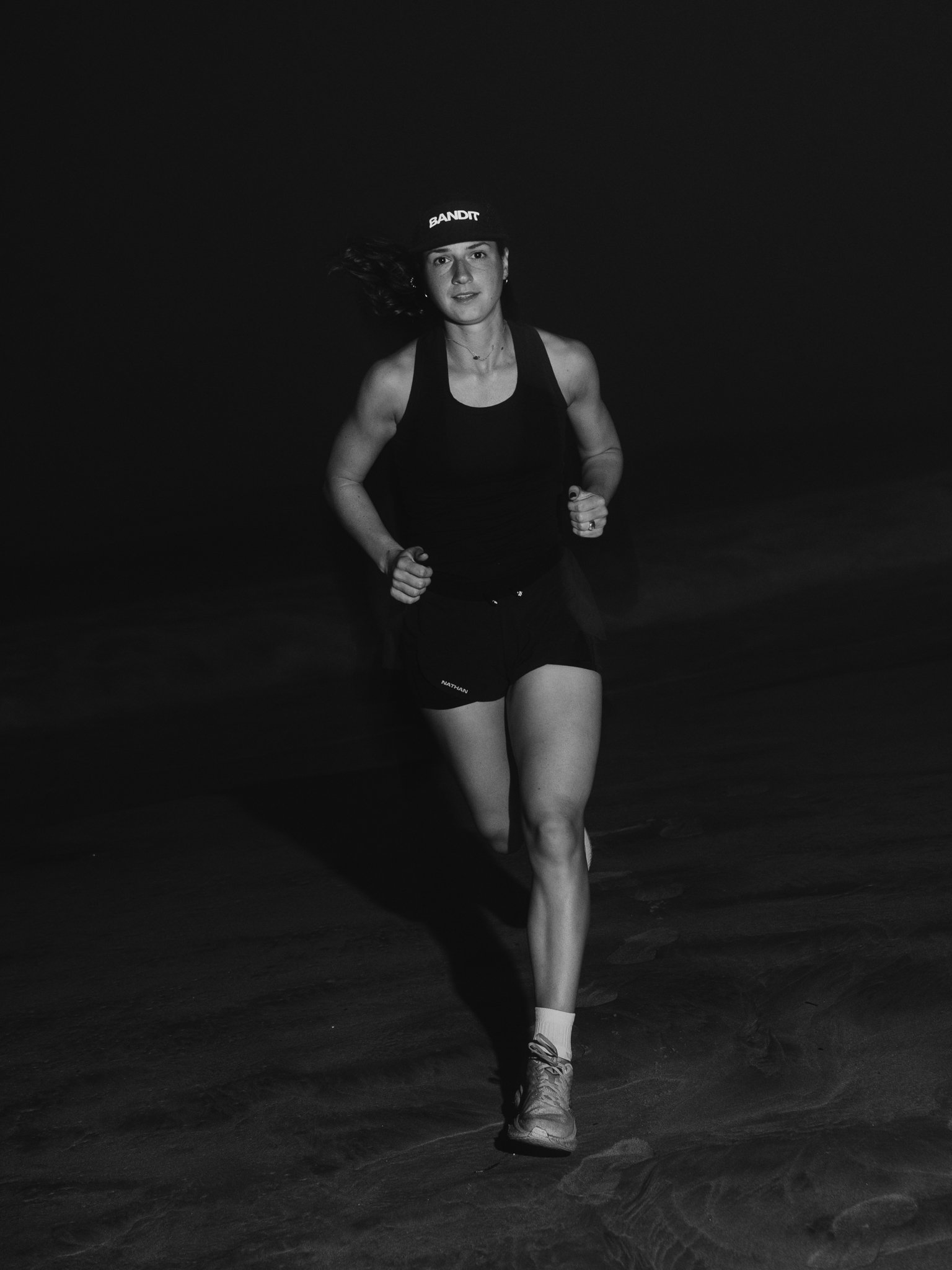 A woman running on a sandy beach at night, wearing a tank top, shorts, sneakers, and a cap that reads 'BANDIT'.
