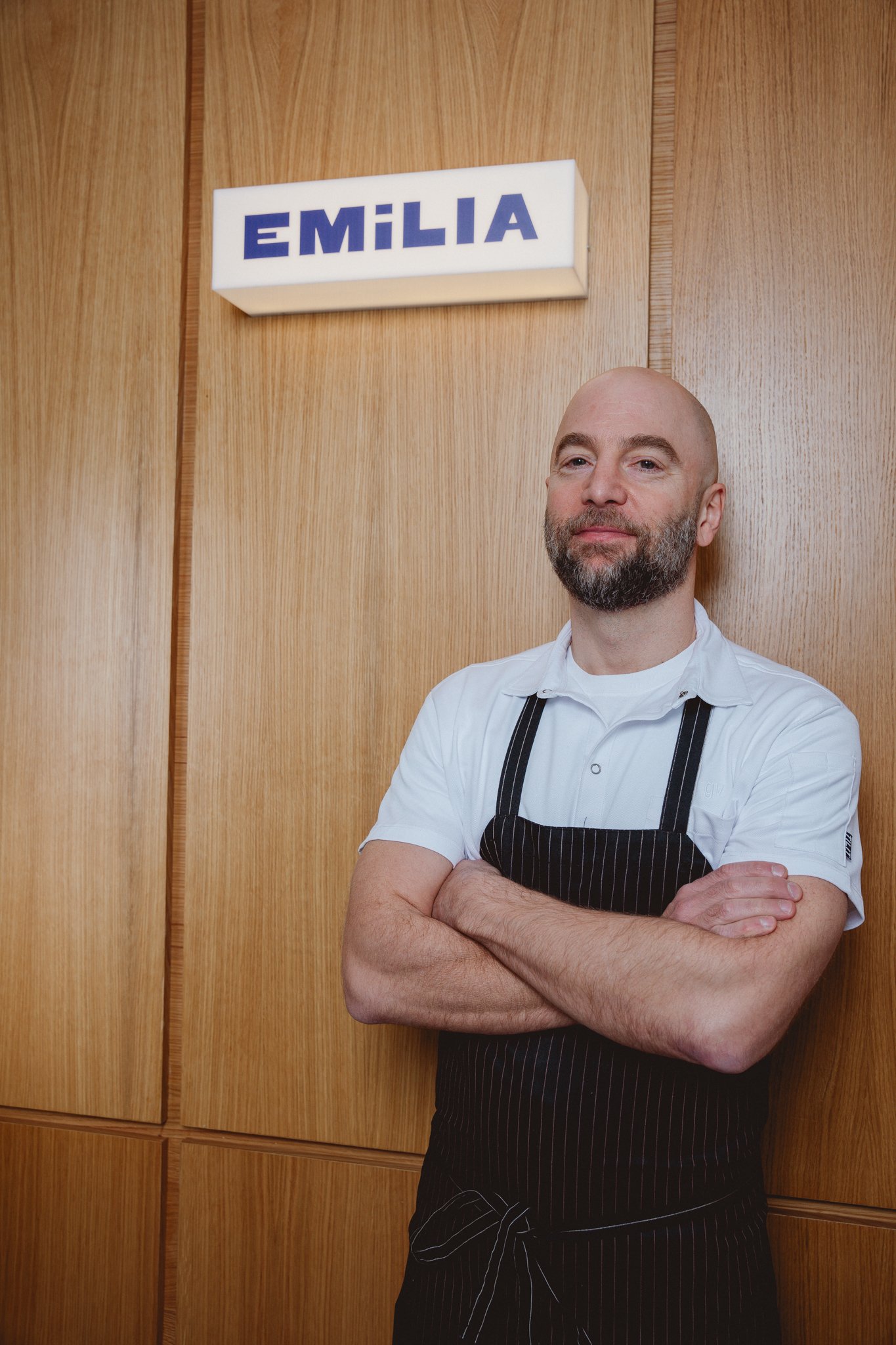 A man with a bald head and gray beard standing with crossed arms in front of wooden cabinets, wearing a white shirt and black apron, with a sign that reads 'EMILIA' mounted on the wall above him.