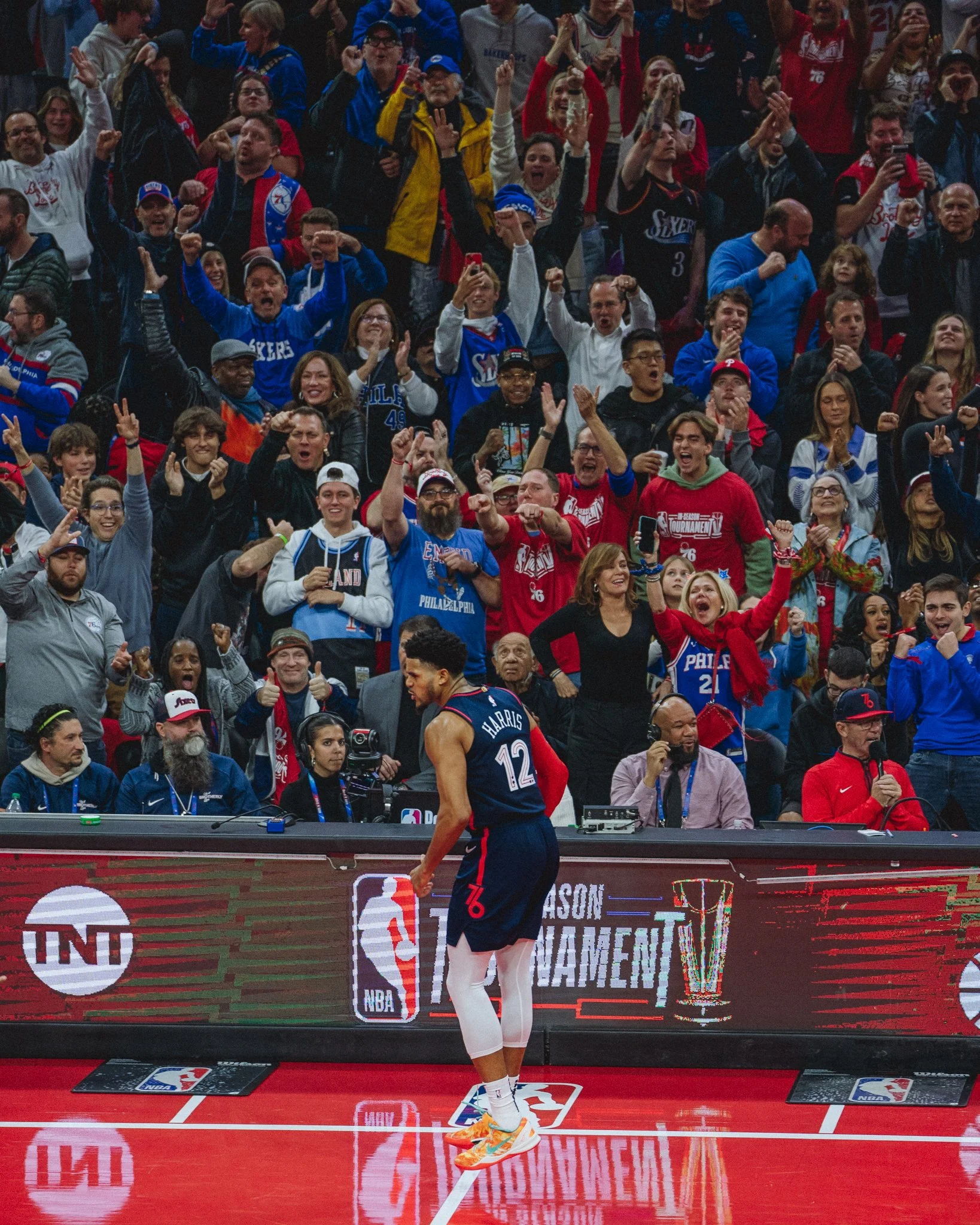 A professional basketball game with a player in the foreground wearing a blue jersey numbered 12, standing on a red court, with a large crowd of fans cheering in the background.