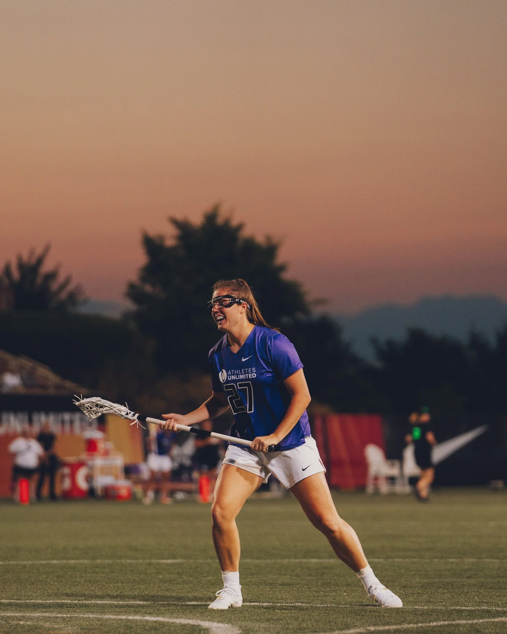 Female lacrosse player in blue and white uniform holding a lacrosse stick on a field during sunset with a tree and crowd in the background.