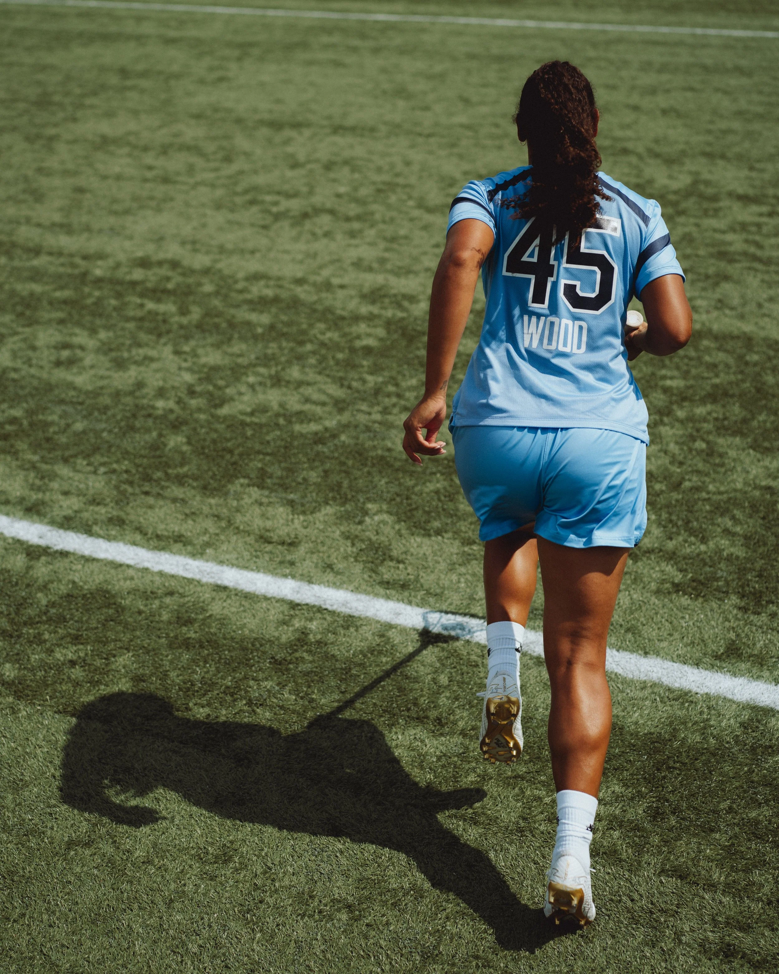 A female soccer player wearing a light blue uniform with the number 45 and the name 'WOOD' on the back, running on a soccer field, casting a long shadow.