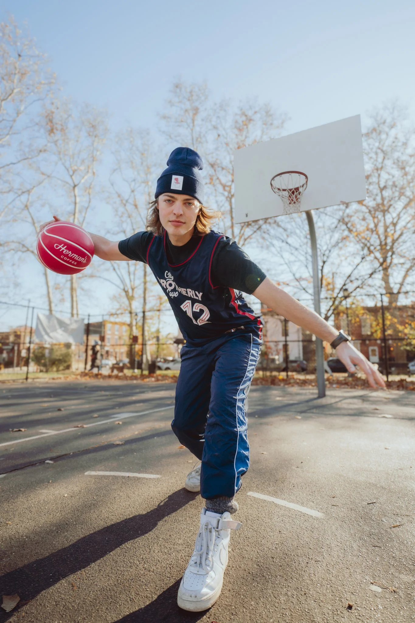 A young man in athletic clothing dribbling a pink basketball on an outdoor basketball court in fall, wearing a dark beanie and white sneakers.