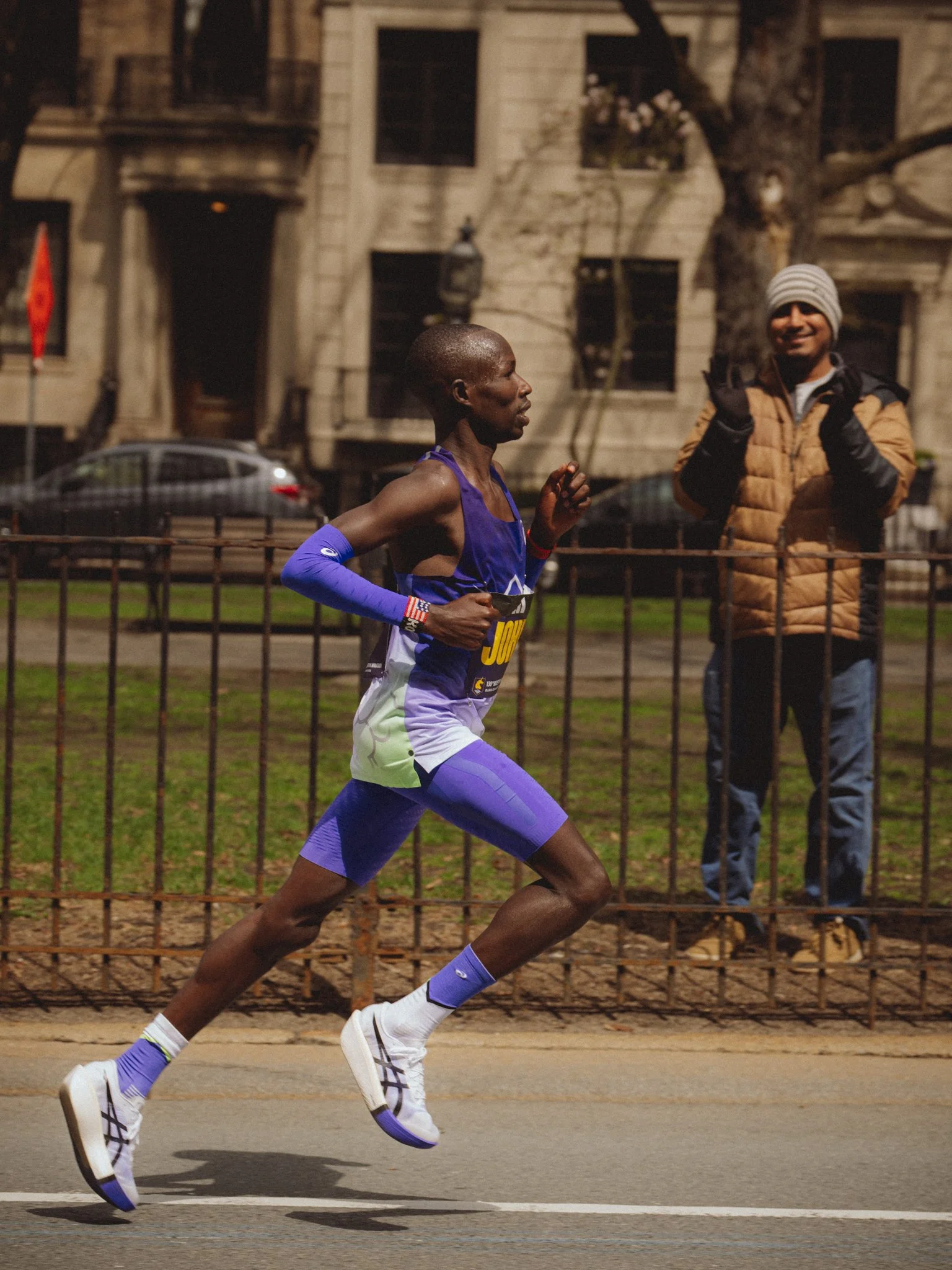 A male runner in a purple and white athletic outfit running on a street, with a man watching behind a metal fence. The background features a building and parked cars, and it appears to be evening or night.