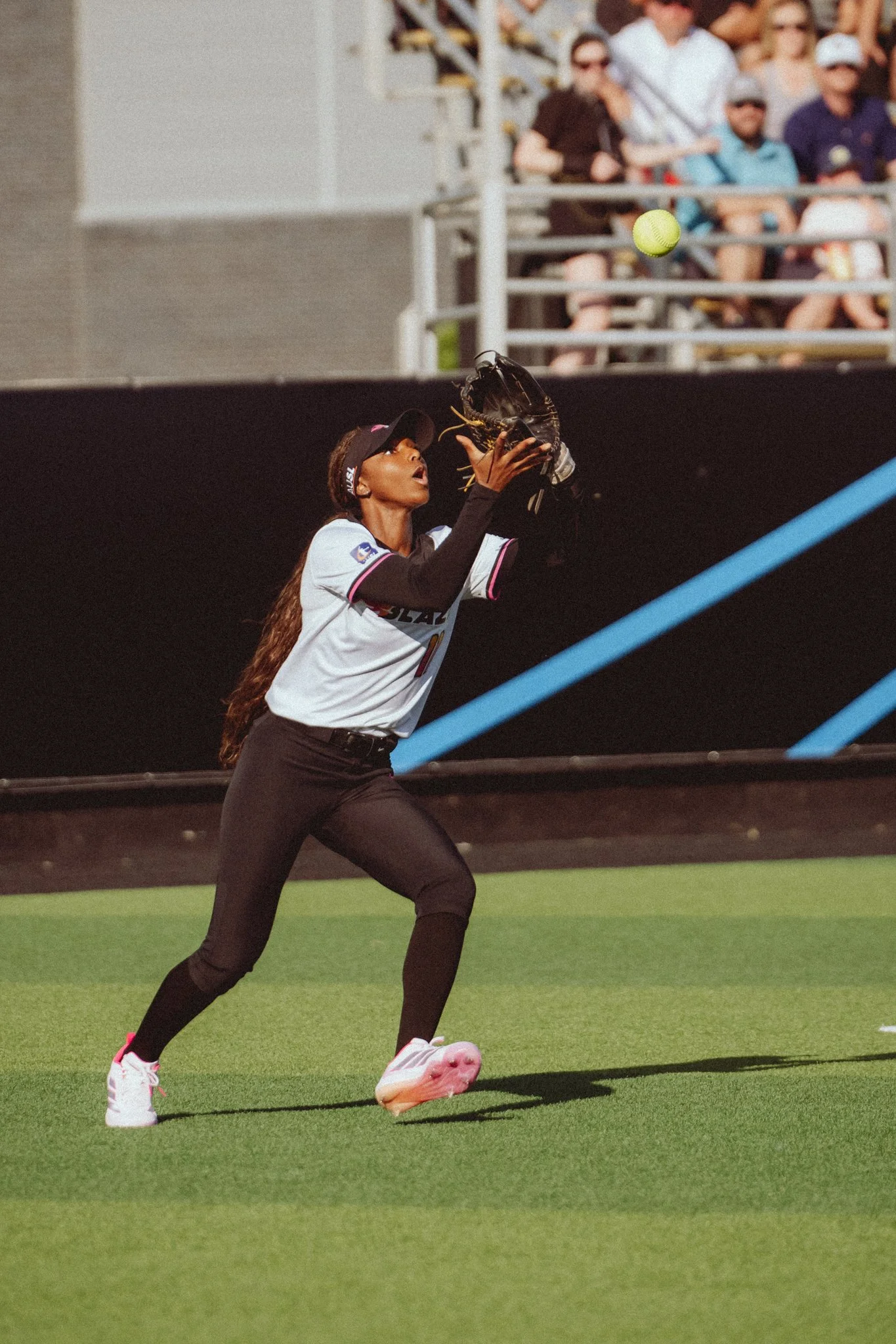 A female tennis player in a black cap and black pants is reaching with her glove to catch a bright yellow tennis ball on a green court, while spectators watch from the stands.
