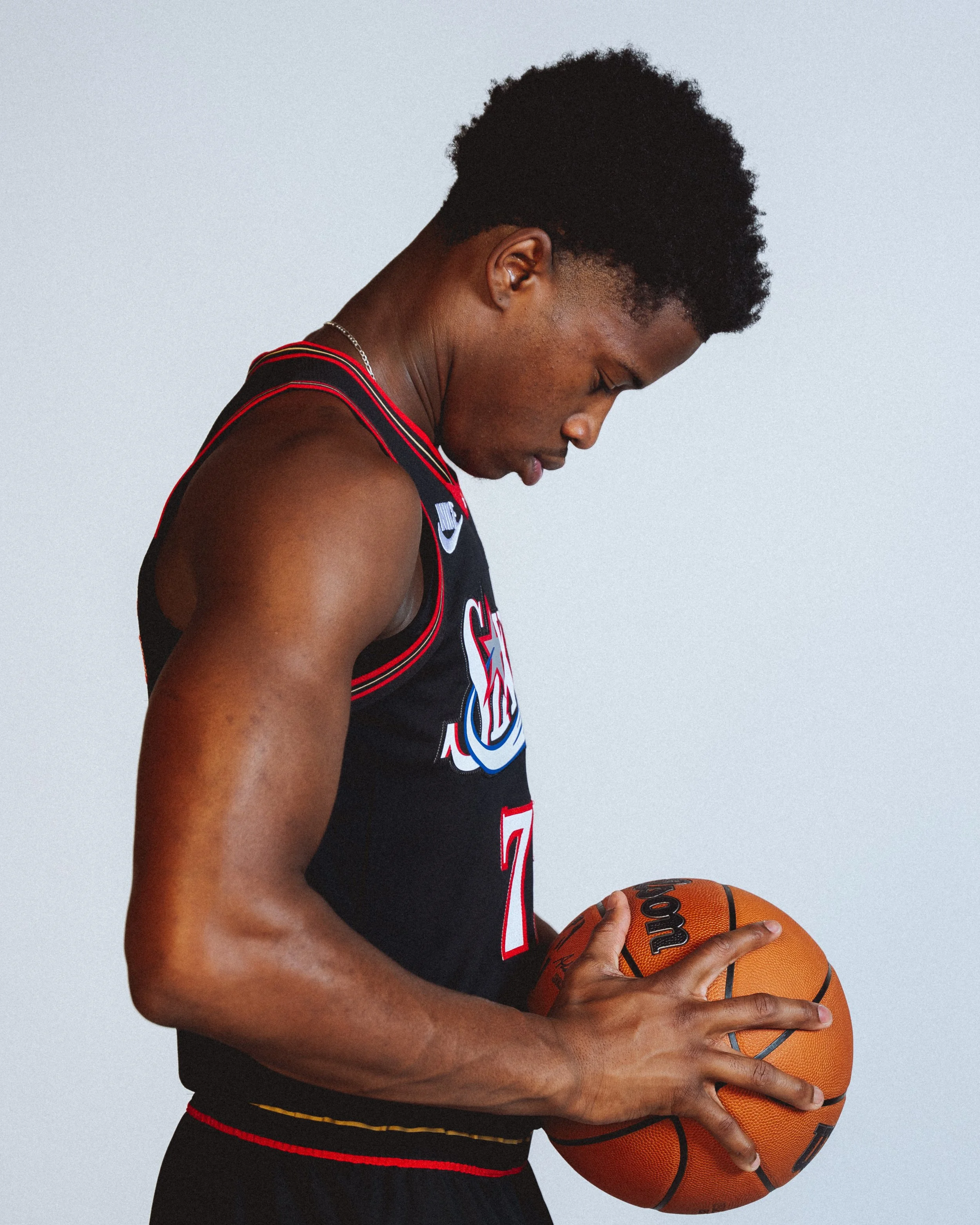 NBA player, VJ Edgecombe wearing a black jersey with red and white accents, holding an orange basketball while looking down, against a plain light-colored background.