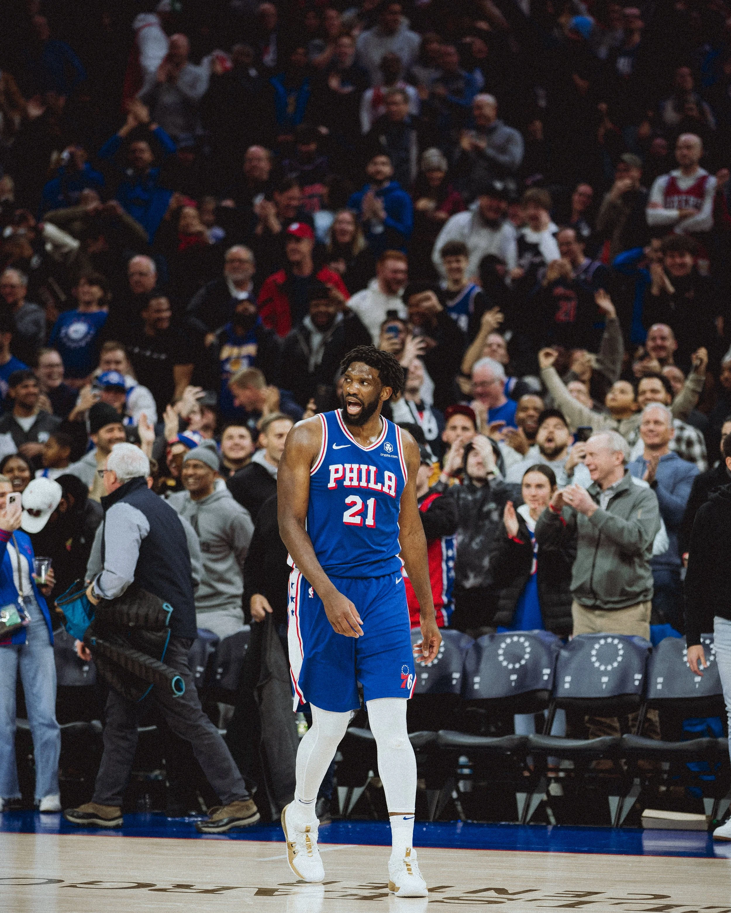 A basketball player from the Philadelphia 76ers, wearing jersey number 21, stands on the court with a crowd of spectators in the background, some clapping and cheering.