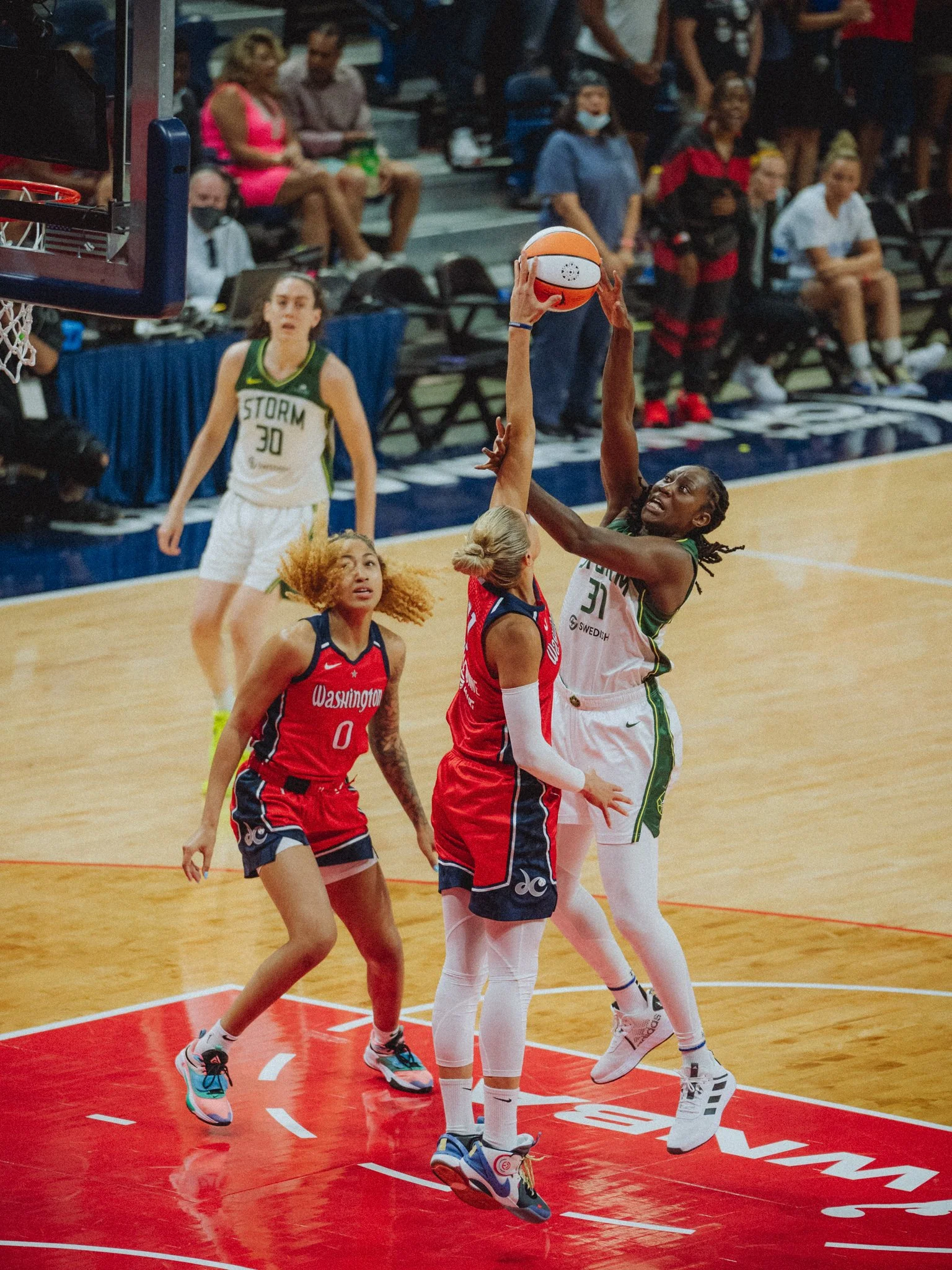 Women’s basketball game with a player in a green and white uniform jumping to shoot a basketball while another player in a red and black uniform attempts to block her, crowd watching in the background.