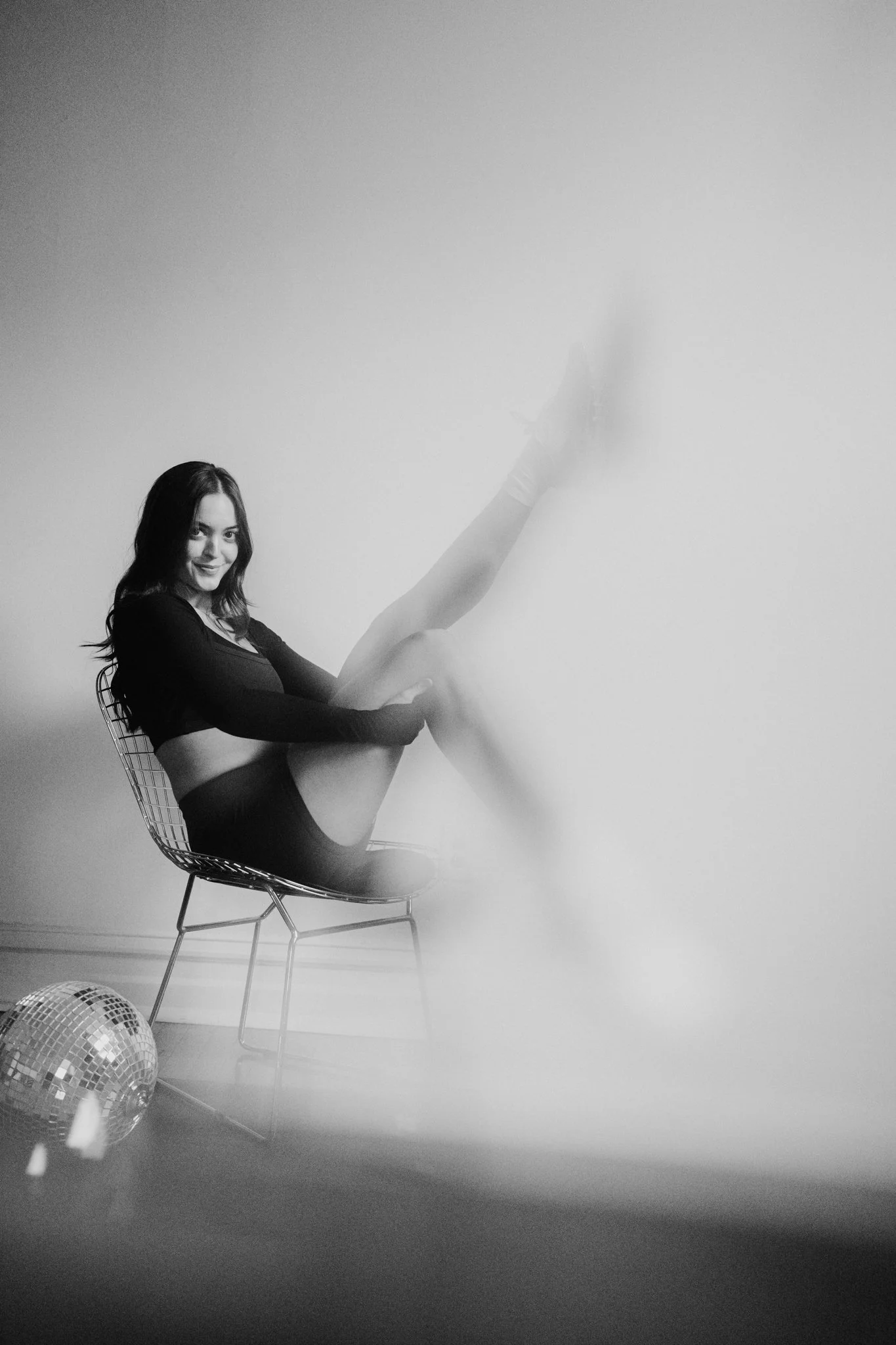 Black and white photo of a woman sitting in a wire chair, smiling, with her hand on her knee. She wears a long-sleeved top and shorts. Two disco balls are on the floor in front of her. The background is a plain, light-colored wall.