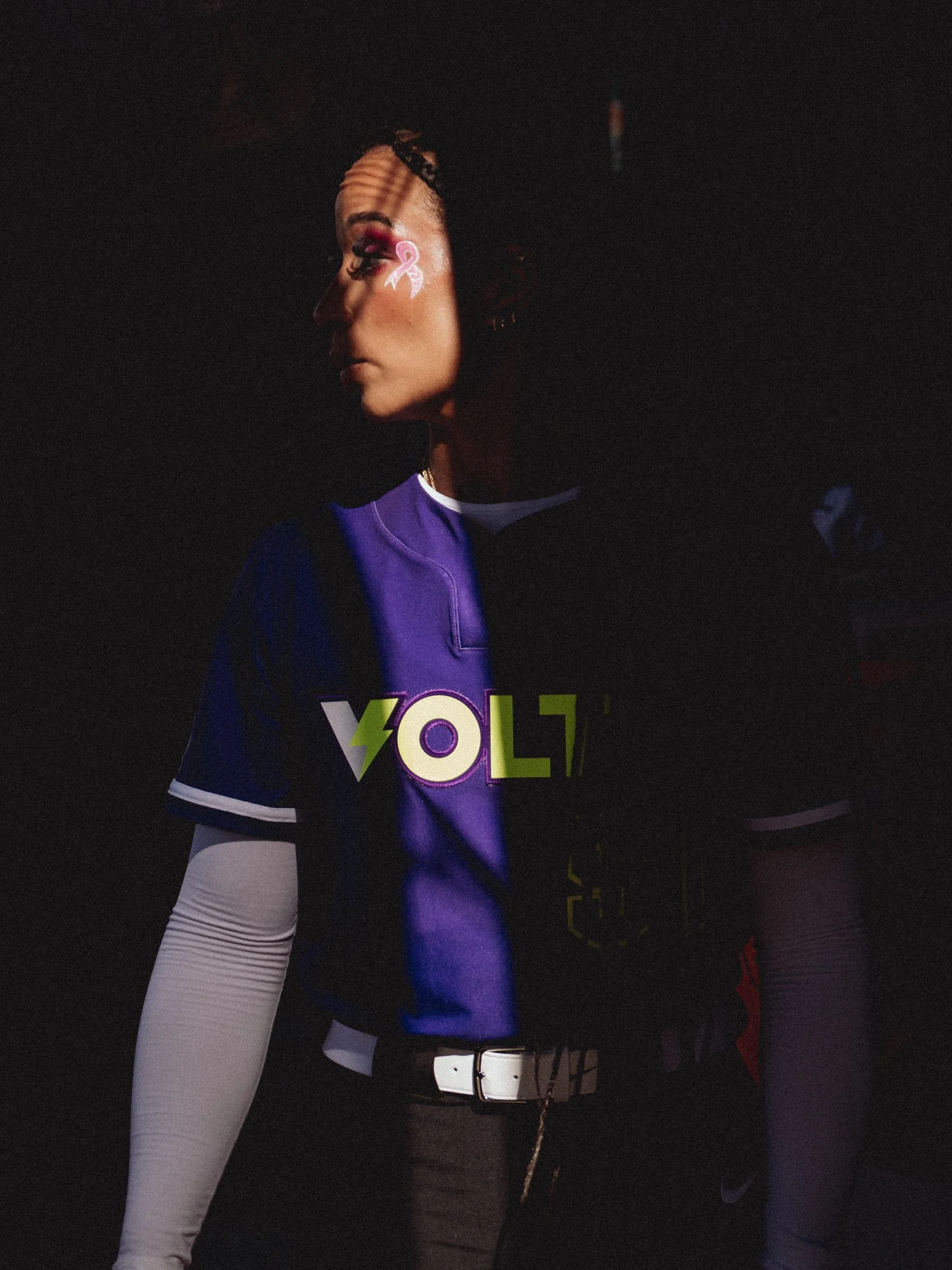 Side profile of a woman with dark curly hair in a dimly lit setting, wearing a purple sports jersey with the word 'VOLT' and a lightning bolt logo, paired with white leggings.