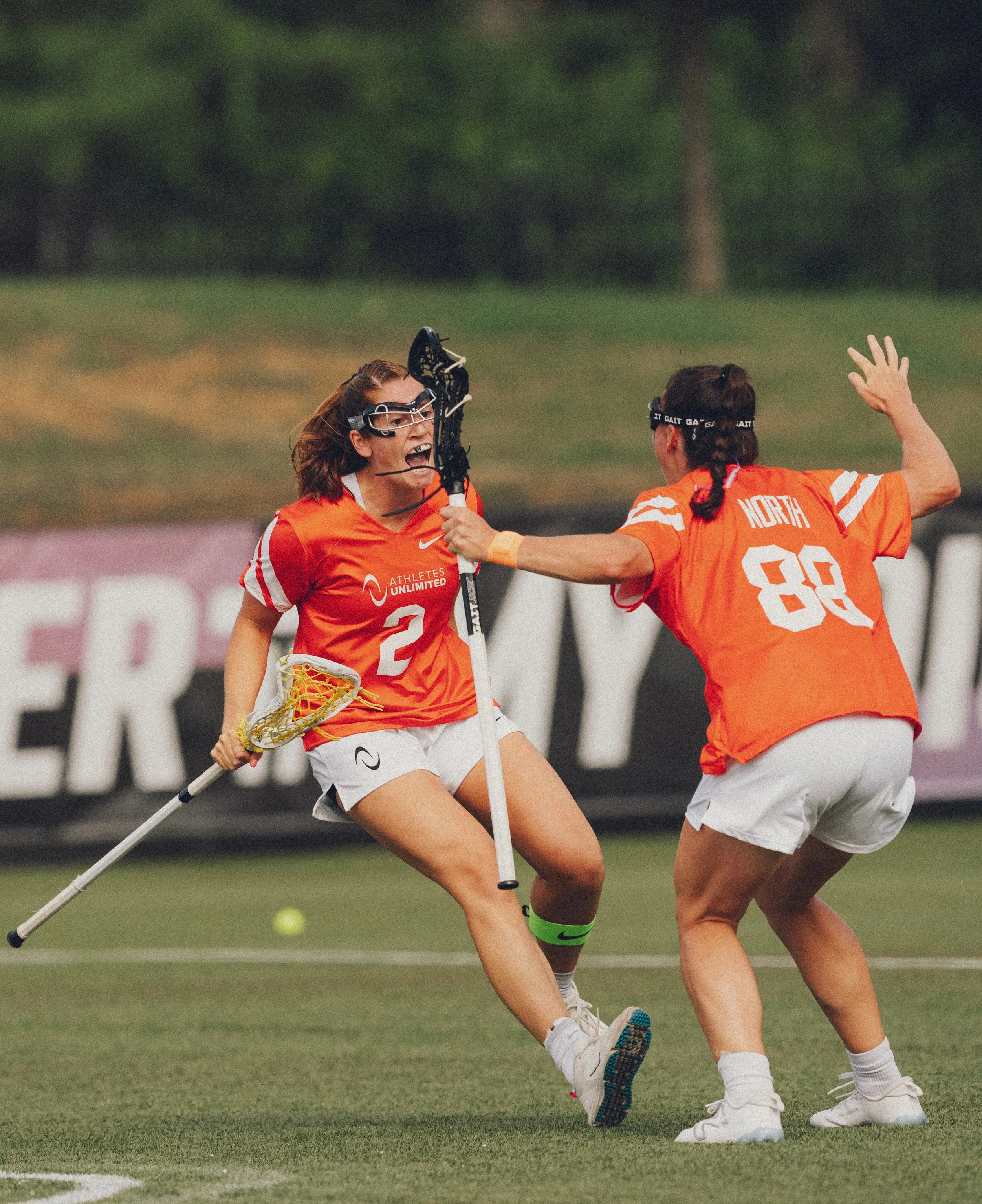Two female field hockey players wearing orange jerseys are competing for the ball on a field, with one player holding a hockey stick and the other raising her hand in a defensive stance.