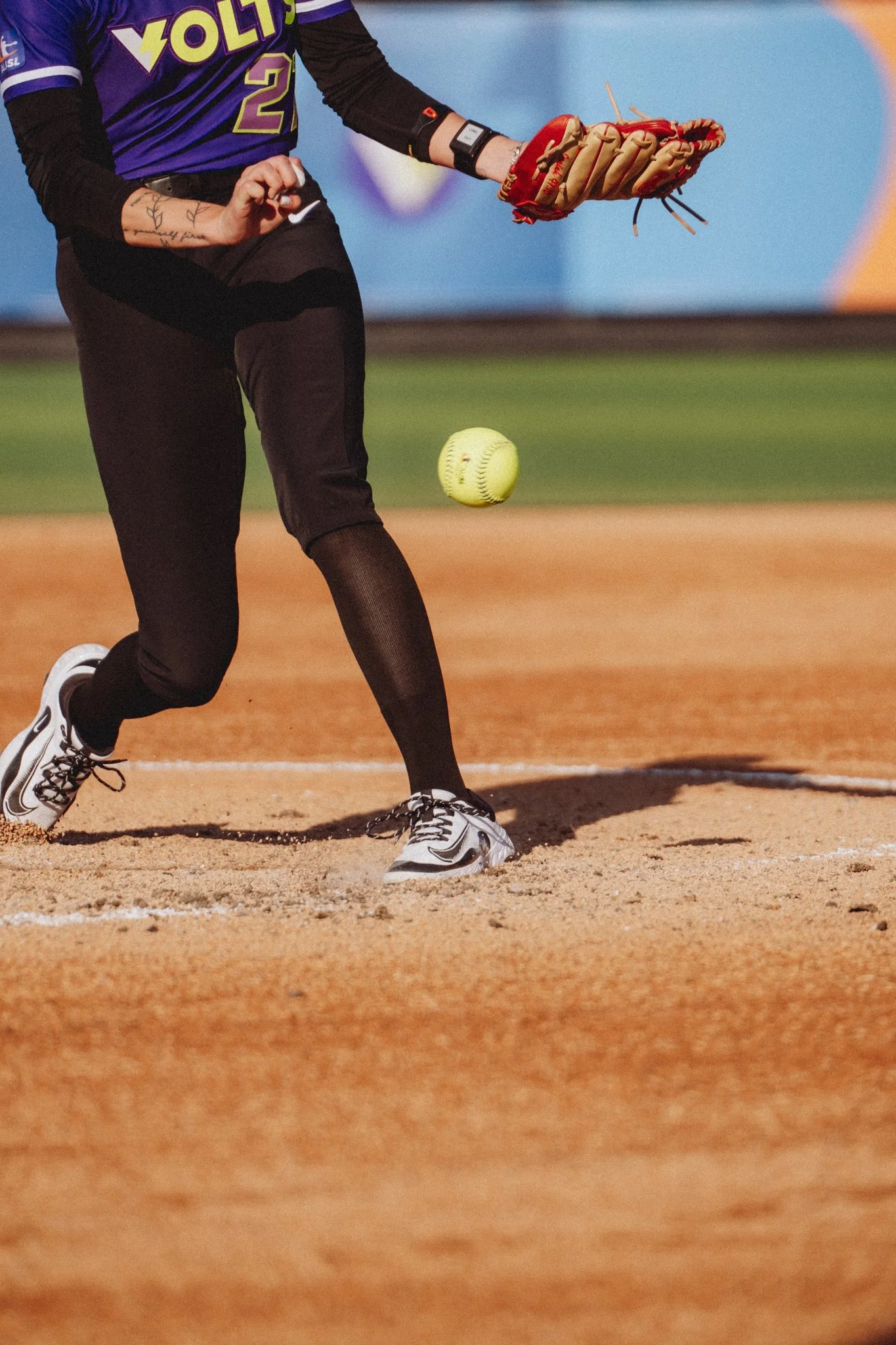 A person wearing a purple sports jersey with the word 'SOLUTION' on it, black pants, and white sneakers, is playing softball on a dirt field. They are holding a red baseball glove and a small cloth in their left hand, with a yellow softball in mid-ai