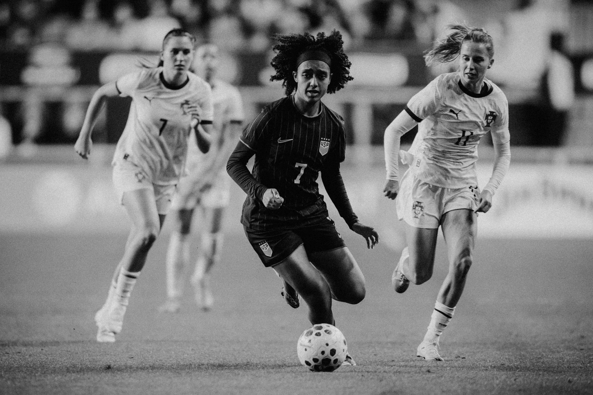 Black and white photo of women playing soccer, with one woman in the center about to kick the ball while others run behind her