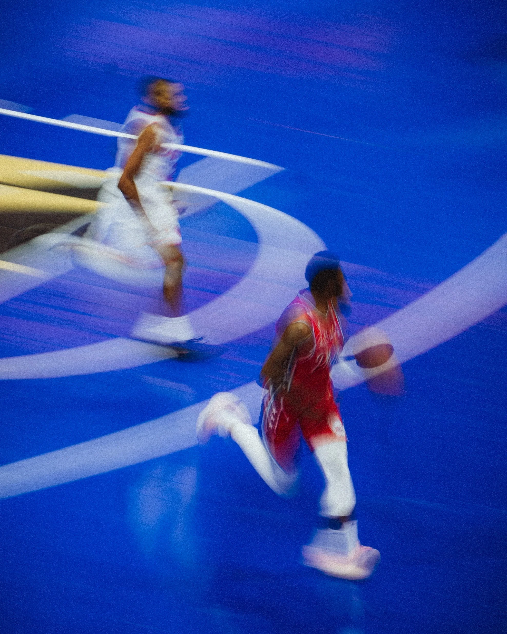 Olympic gymnast executing a routine on the balance beam in a large indoor gymnasium with blue flooring, blurred motion indicating movement.