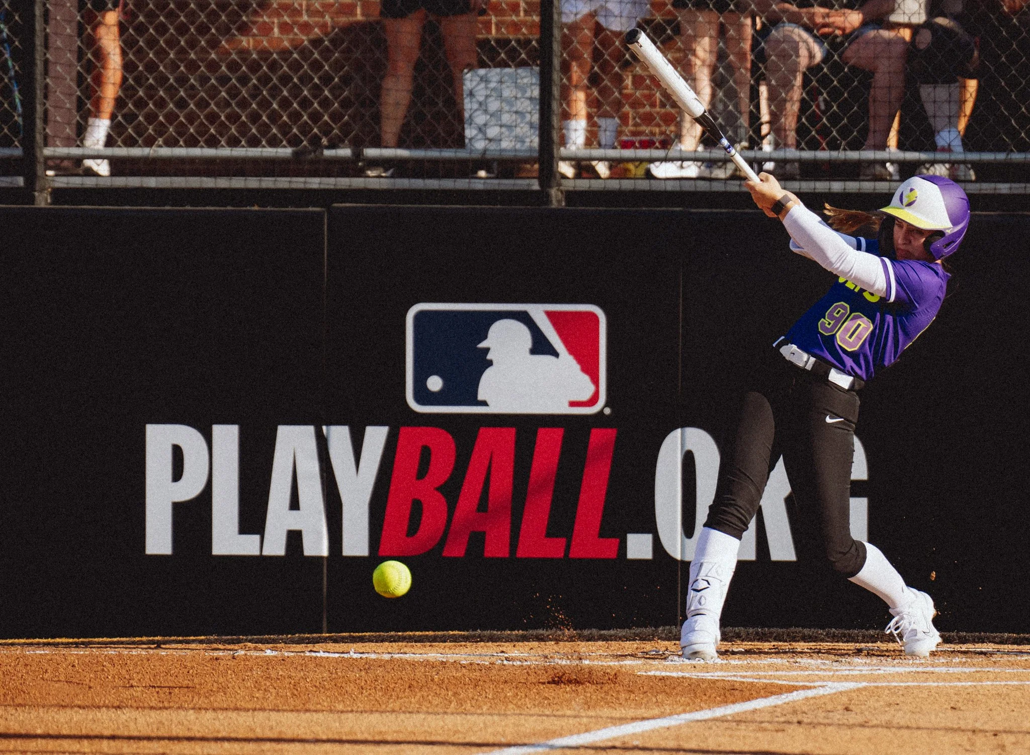 A female softball player swinging a bat at a ball during a game, with a black fence and spectators in the background, and the MLB Play Ball logo on the outfield wall.