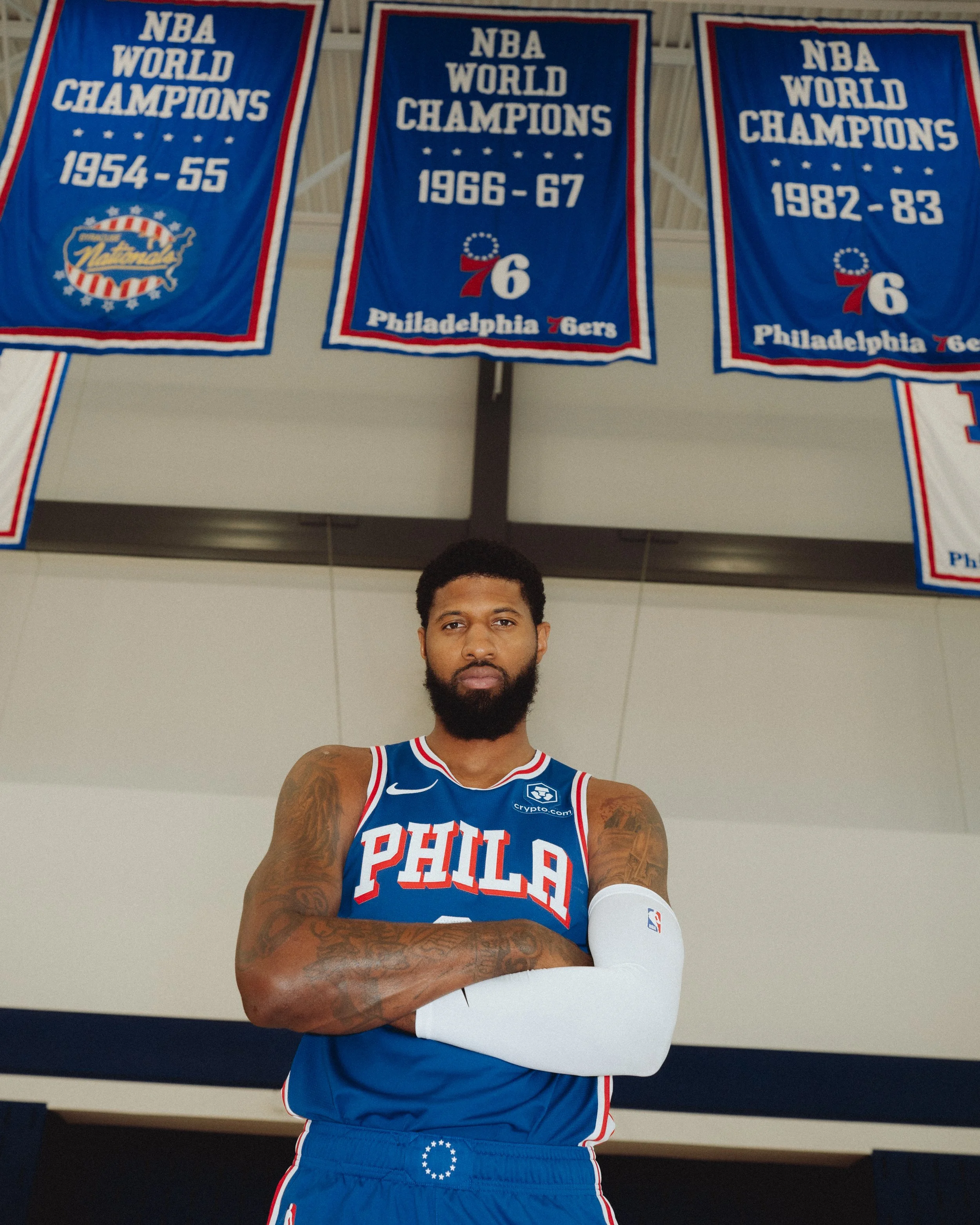 A male basketball player wearing a Philadelphia 76ers jersey and a white arm sleeve, standing in a gym with basketball banners hanging in the background.