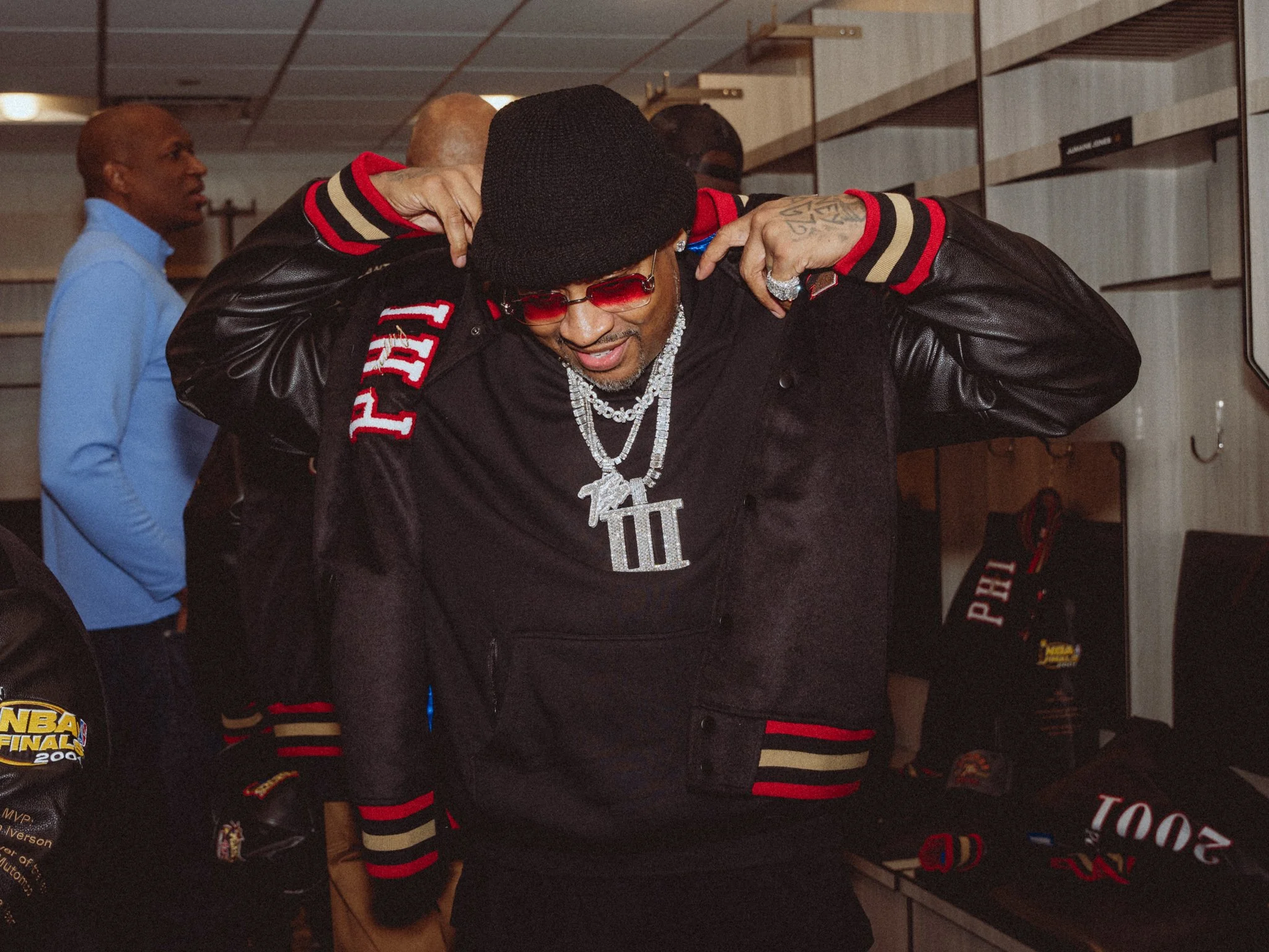 A man wearing a black hat, red sunglasses, and jewelry embraces another person in an indoor setting, with a man in a blue shirt visible in the background.