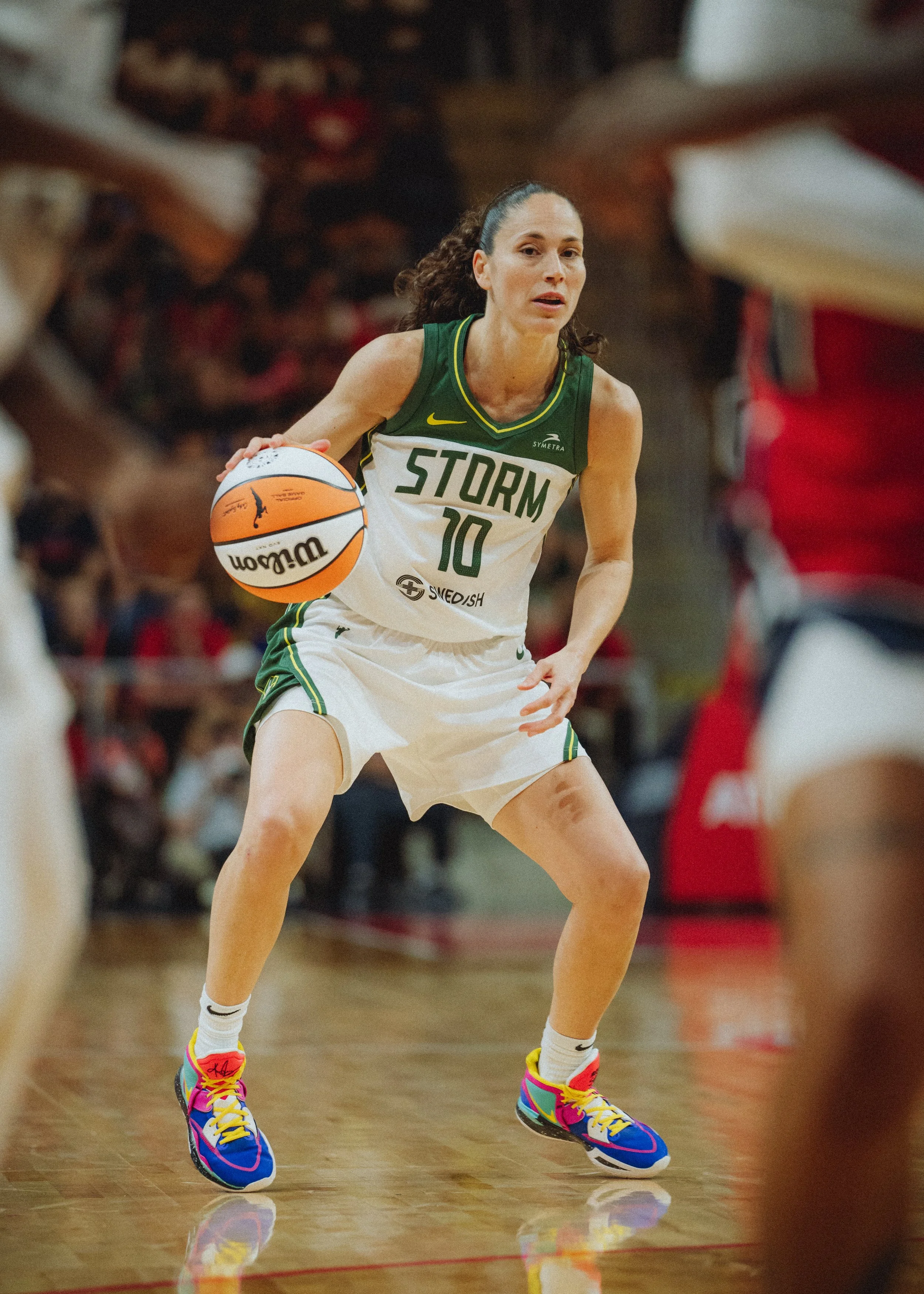 A female basketball player wearing a green and white uniform with the number 10 dribbling a basketball on a court. She has dark curly hair tied back and colorful sneakers. The court has a wooden floor with red lines, and there are spectators blurred 