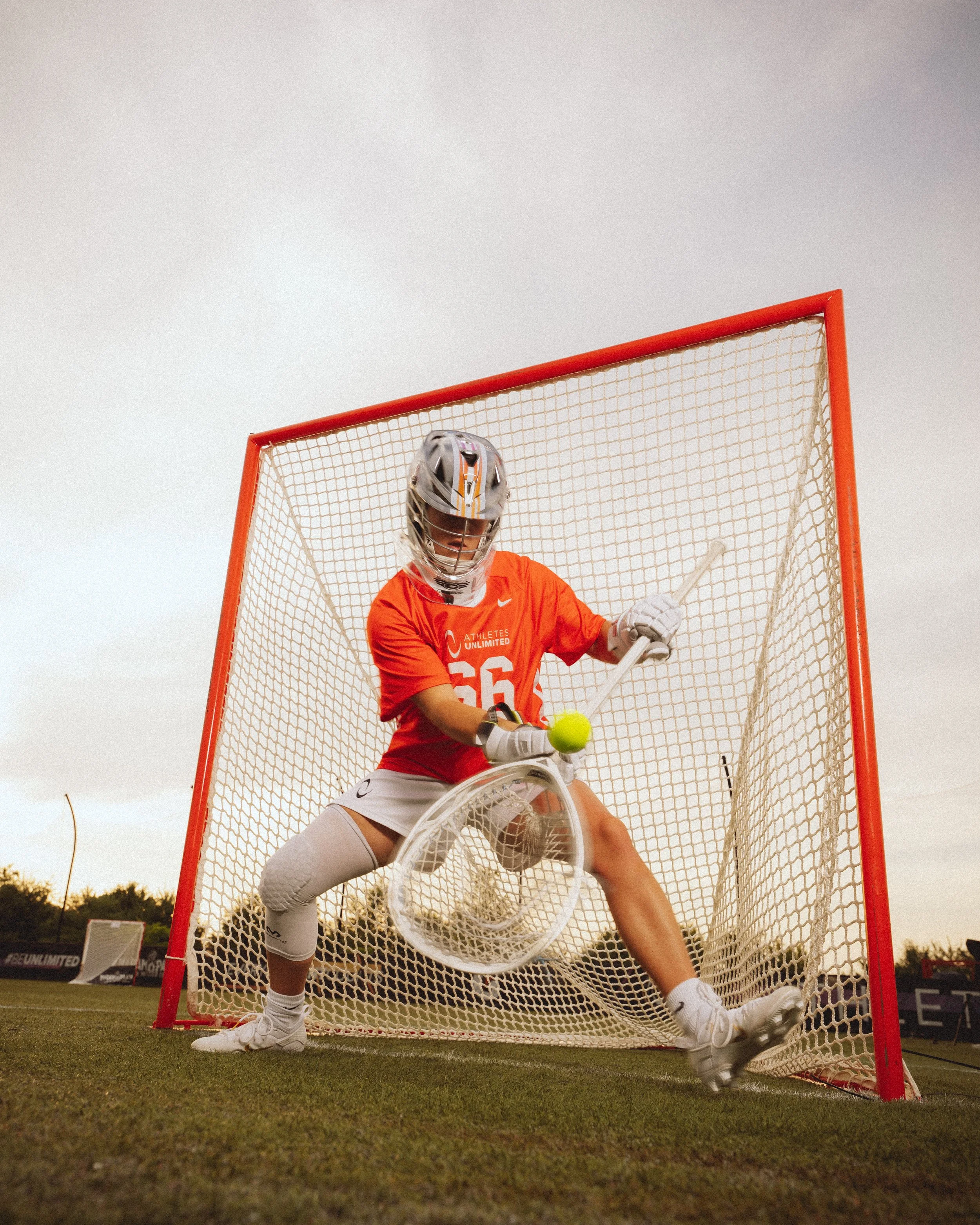 A hockey goalie in orange sportswear stands in front of the goal, holding a stick and catching a yellow hockey ball with their glove during a game on an outdoor field.
