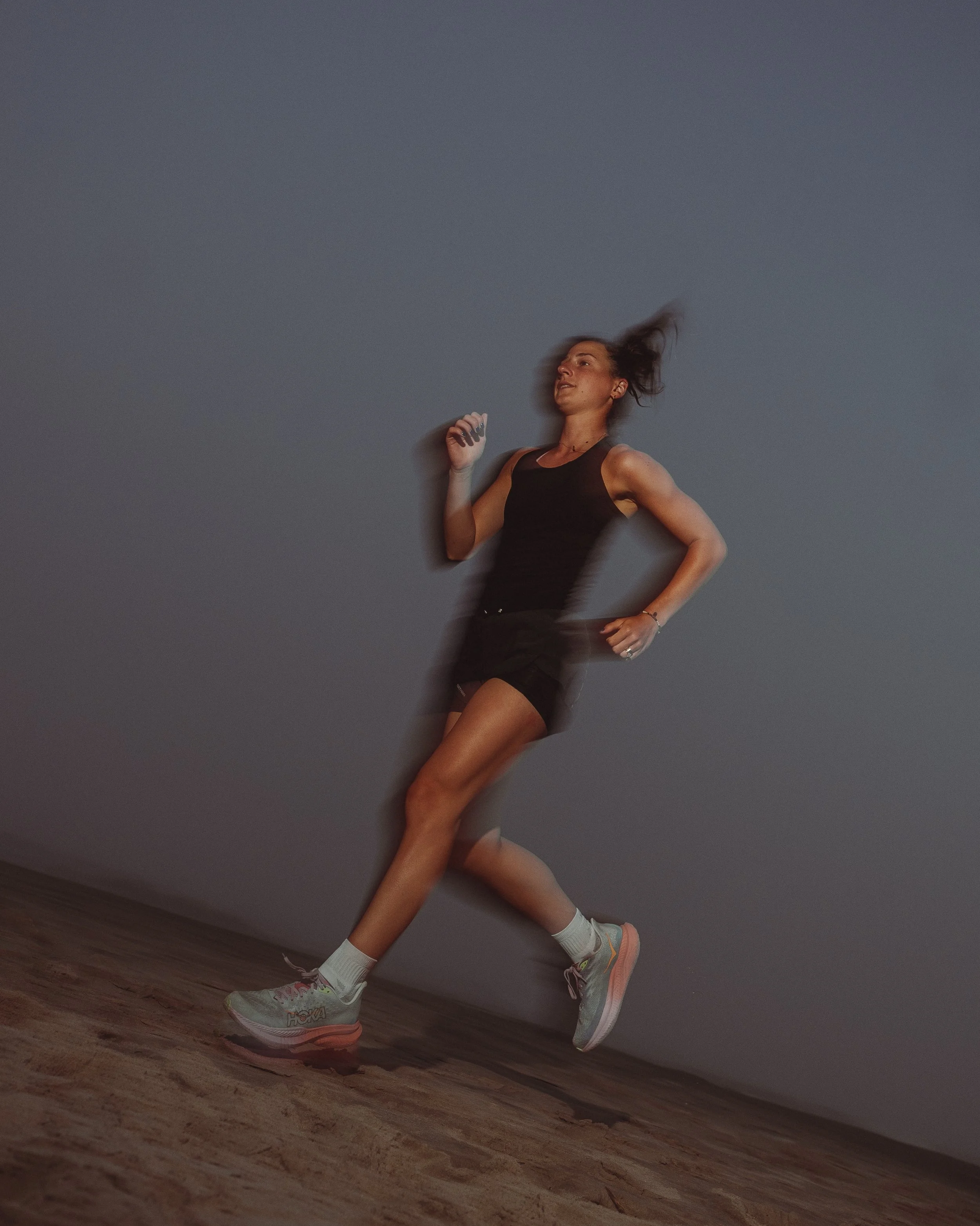 A woman running indoors in front of a gray wall, wearing athletic clothing including a black tank top, shorts, and running shoes. Her hair is in motion and her face shows effort.