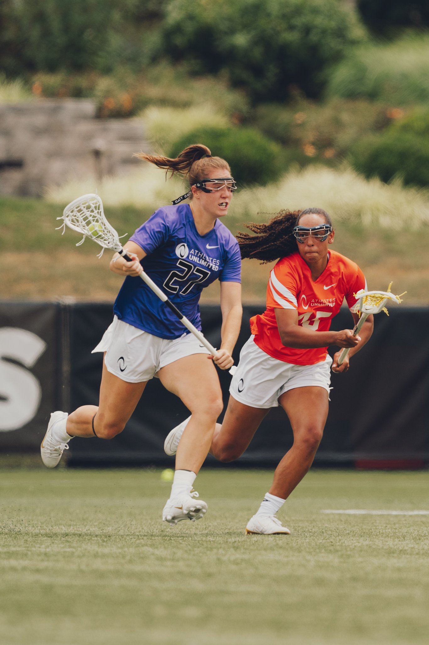 Two female lacrosse players running on a field, one in a blue jersey and the other in a red jersey, both wearing safety goggles and holding lacrosse sticks.